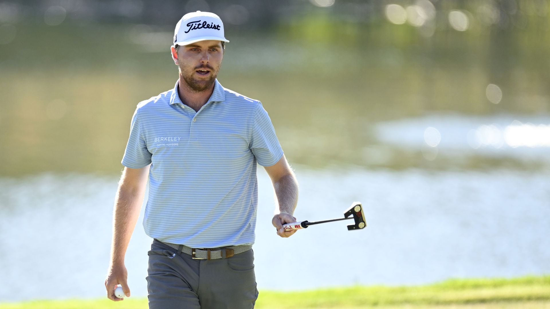 Male golfer in light blue striped shirt and white Titleist cap walking on a green golf course near water, holding a golf ball and a putter.