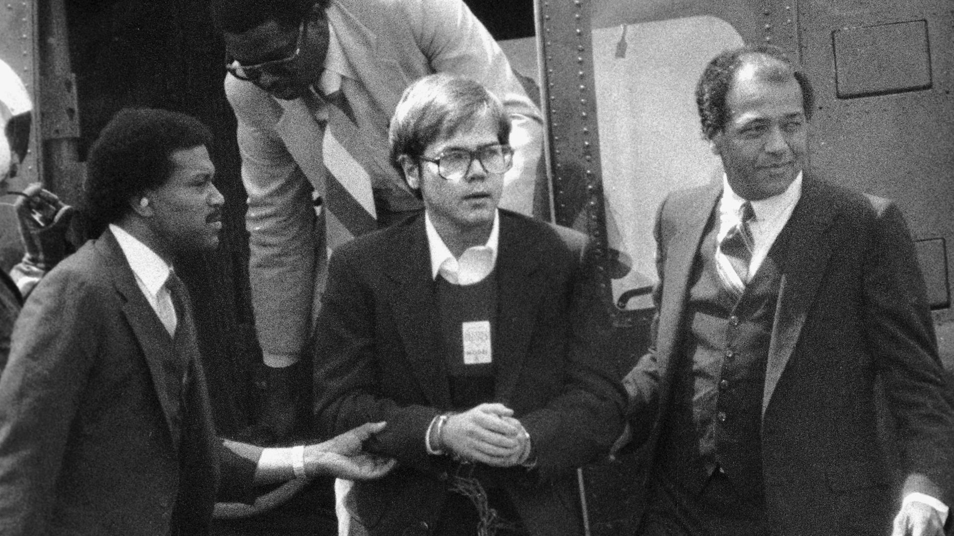 Photo of a man handcuffed with one man holding each arm as they exit a helicopter