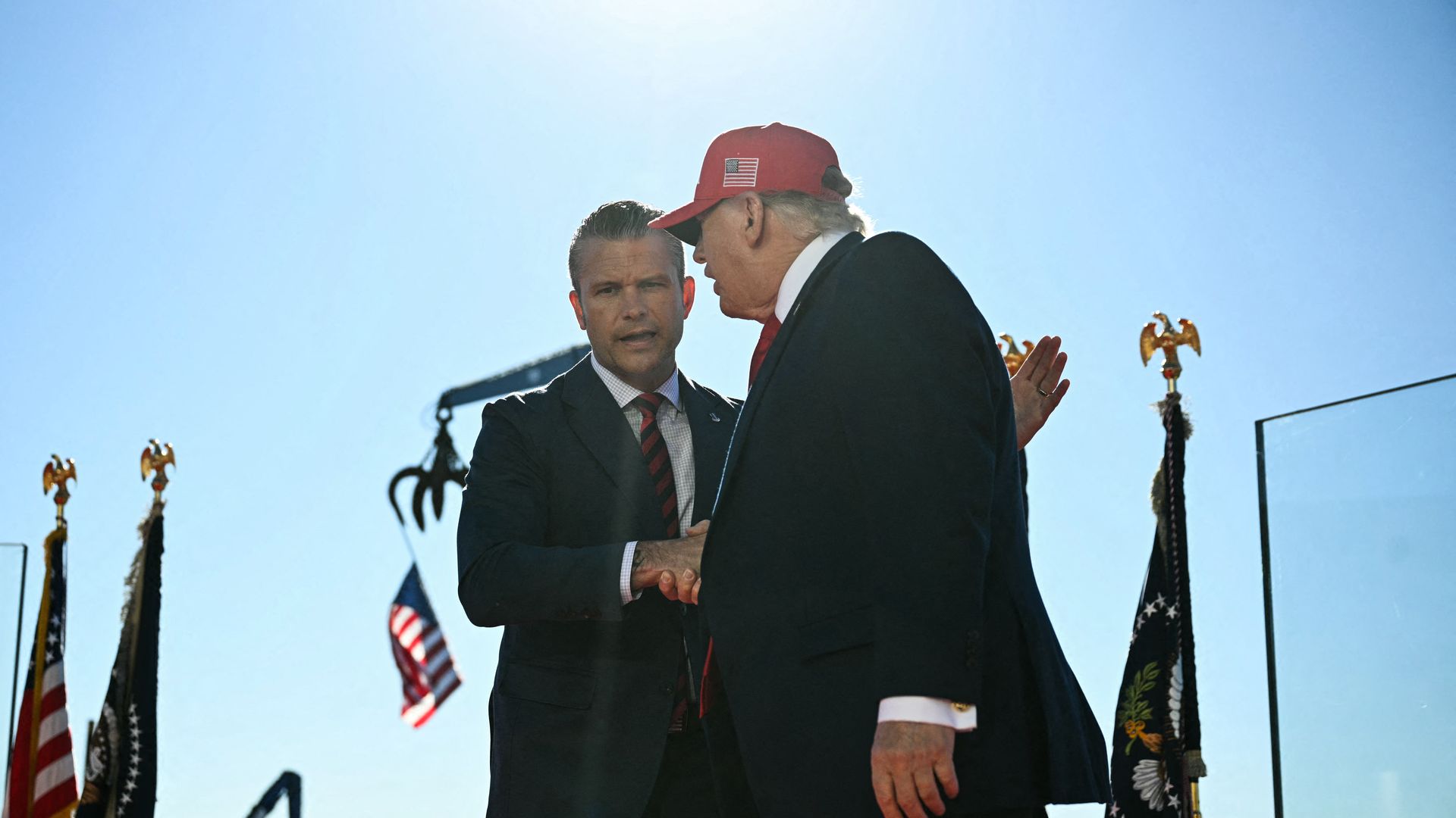 Trump — wearing a red hat and a suit — shakes hands with Pete Hegseth — who is wearing a suit and a red striped tie — while the two men stand outside near flags.
