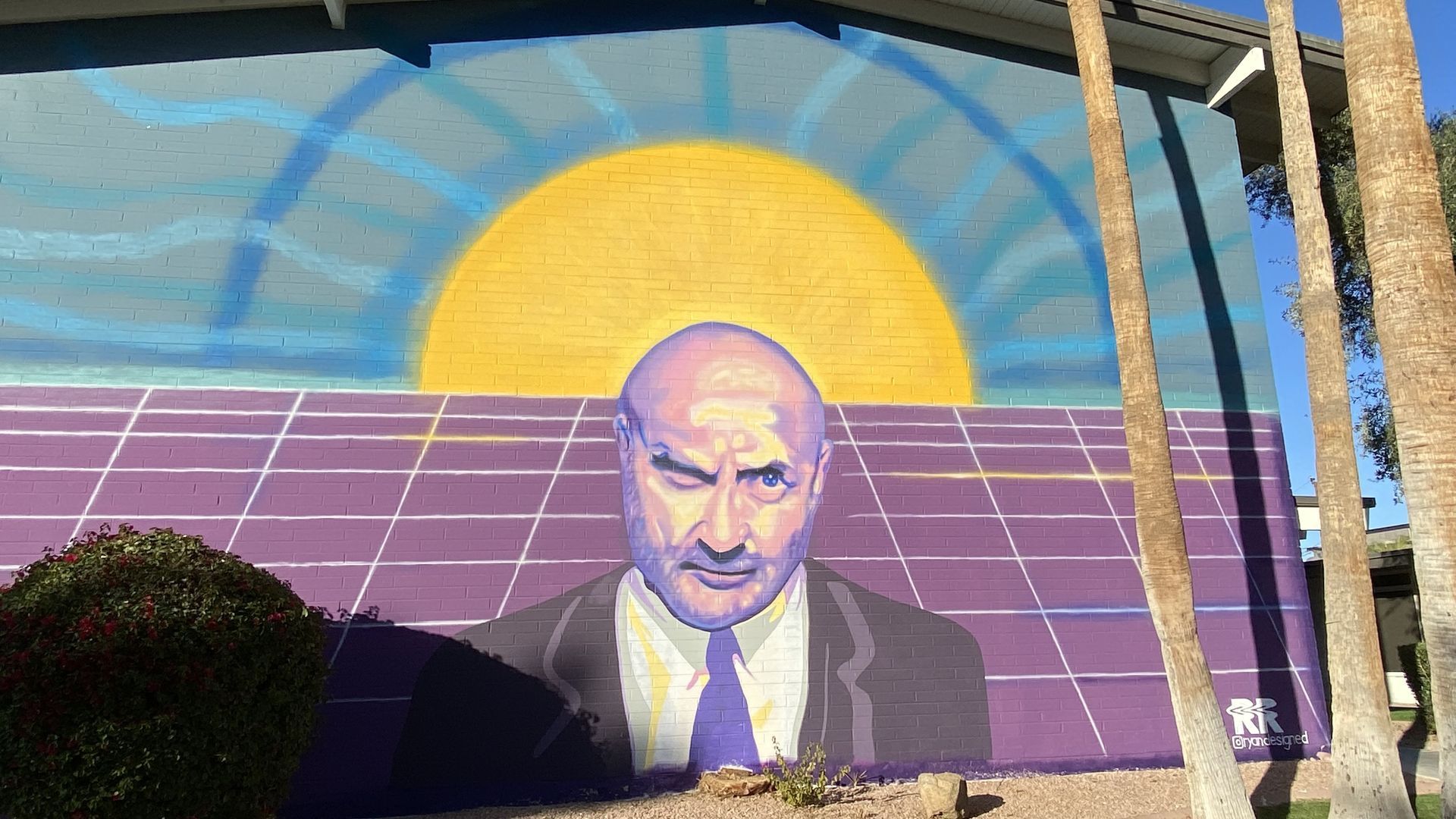 A mural depicting Phil Collins in a suit against a purple wall, with a yellow sun rising over it, surrounded by a blue halo. 