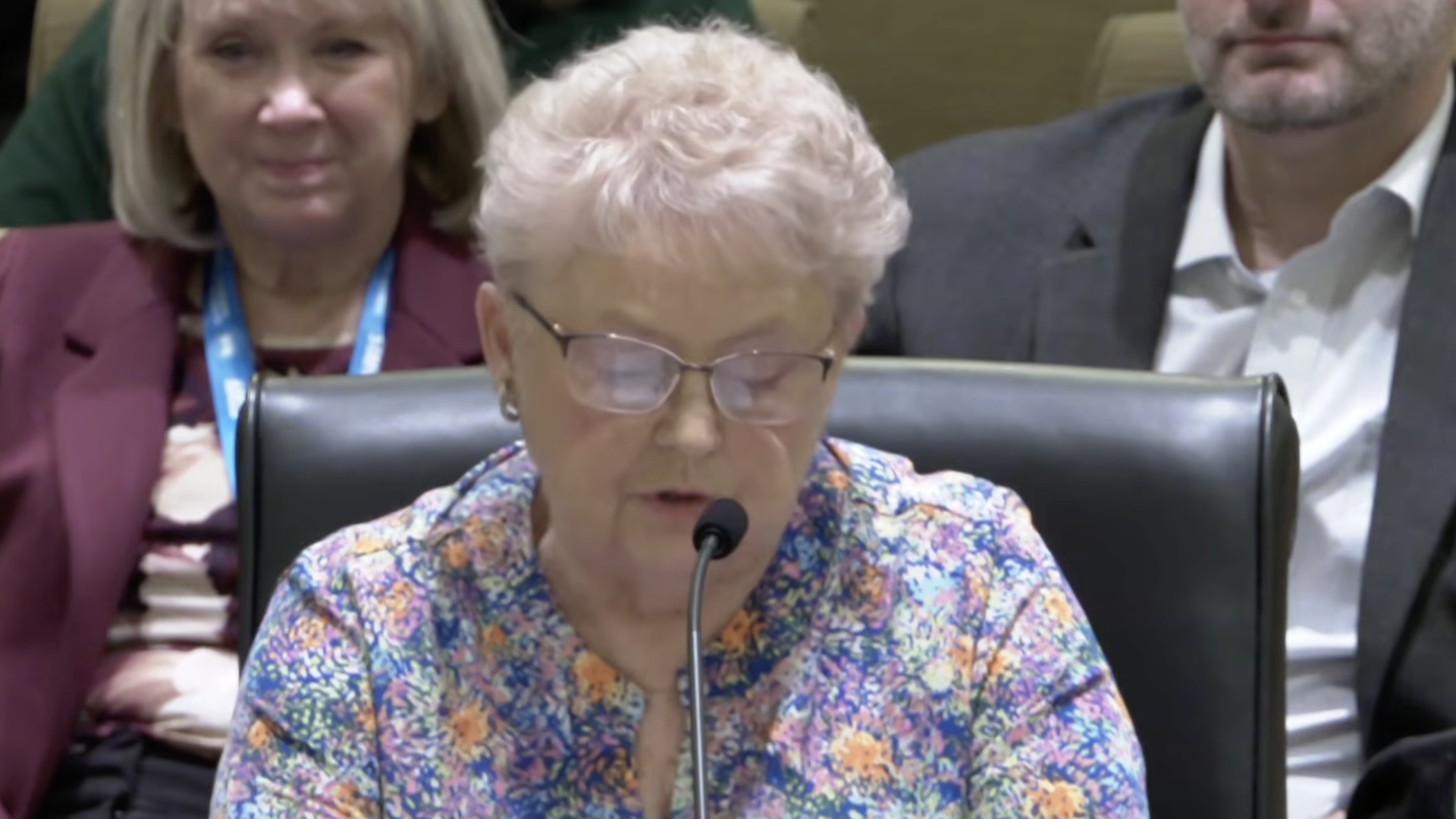 An older woman with short white hair and glasses reads from a paper while speaking into a microphone at a panel; she wears a bright floral blouse, with two people in suits seated behind her.