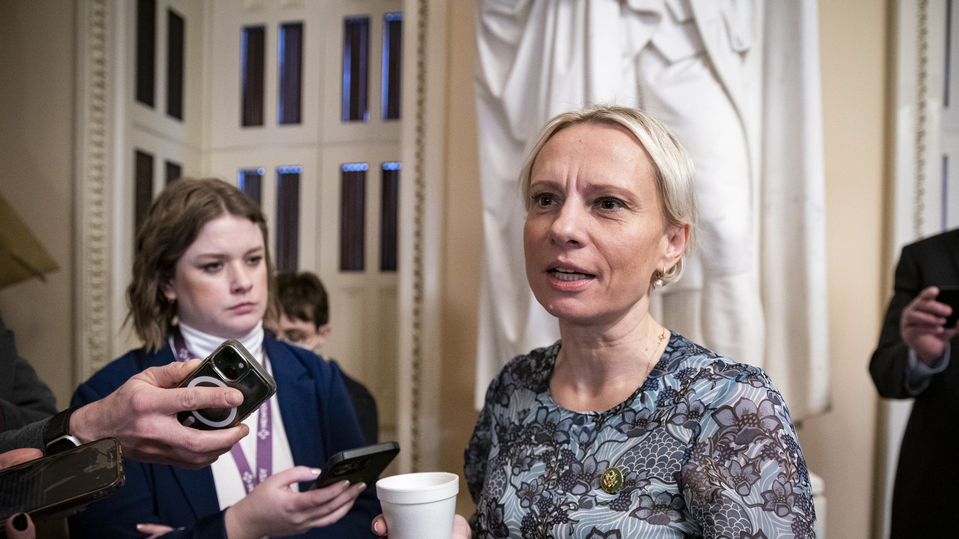 Rep. Victoria Spartz, wearing a blue dress and holding a styrofoam cup, speaks to reporters in front of a statue at the Capitol.