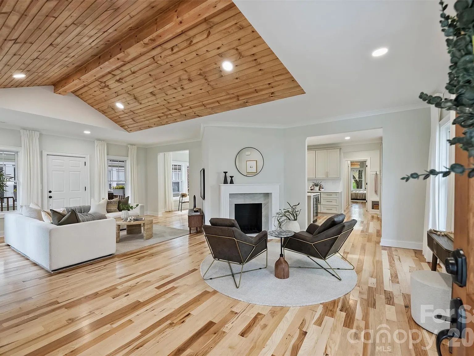 Open living room with a vaulted wooden ceiling, light wood floors, and white walls. A white sofa and two black chairs surround a round table on a gray rug by a fireplace with a round mirror.