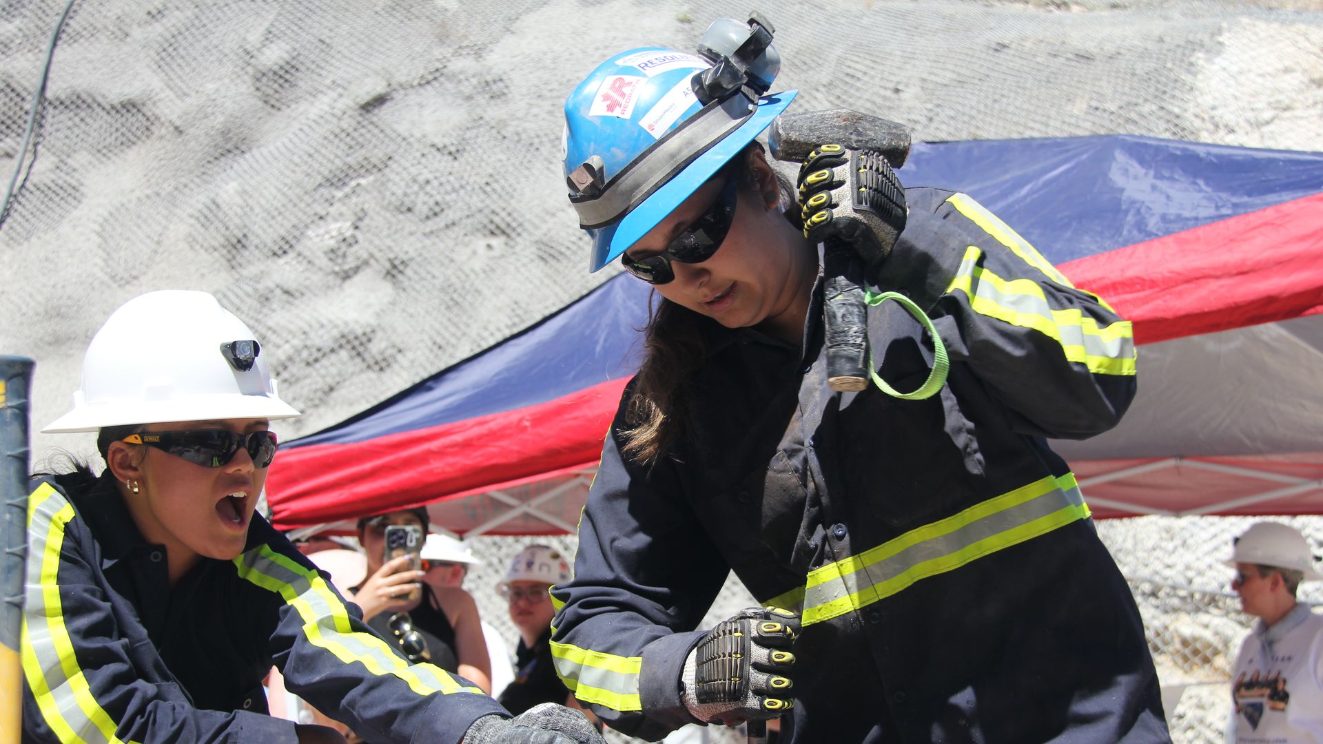 Two women in mining helmets and sunglasses, wearing what looks like firefighting gear, work on a concrete surface with a large hammer, while onlookers watch under a tent.