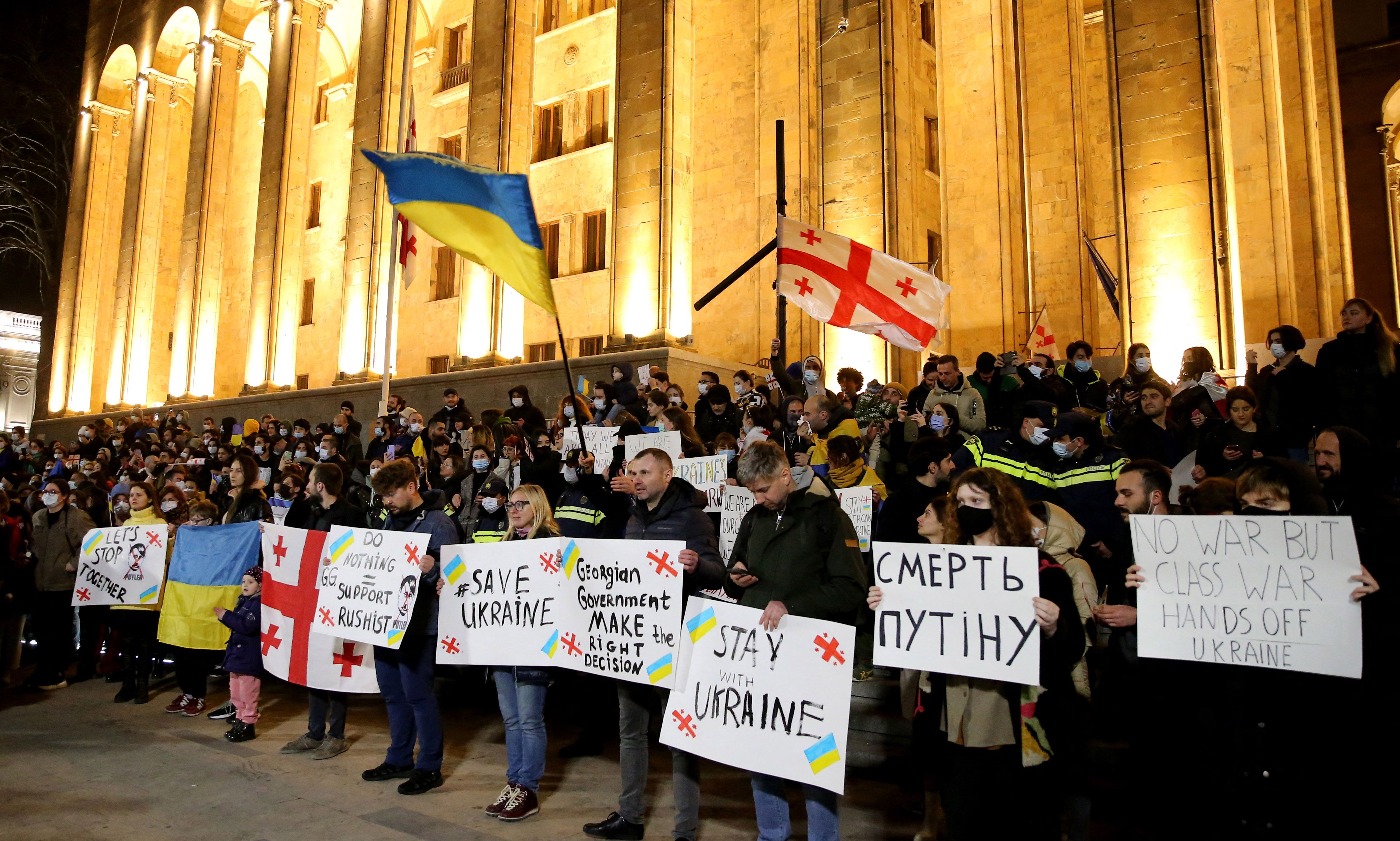 Photo of people holding signs and waving flags in support of Ukraine at night in front of a large lit-up building