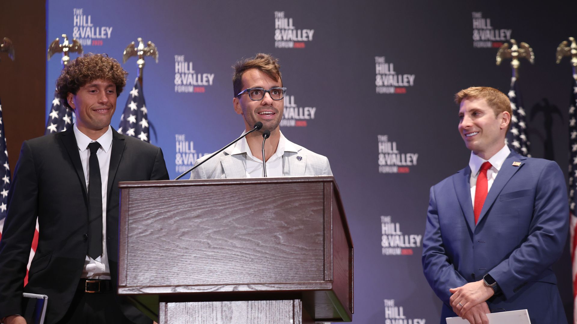 Three men stand on stage. Two are in suits, one black and one blue. The other man is in a collared white shirt. There is podium as well as American flags.
