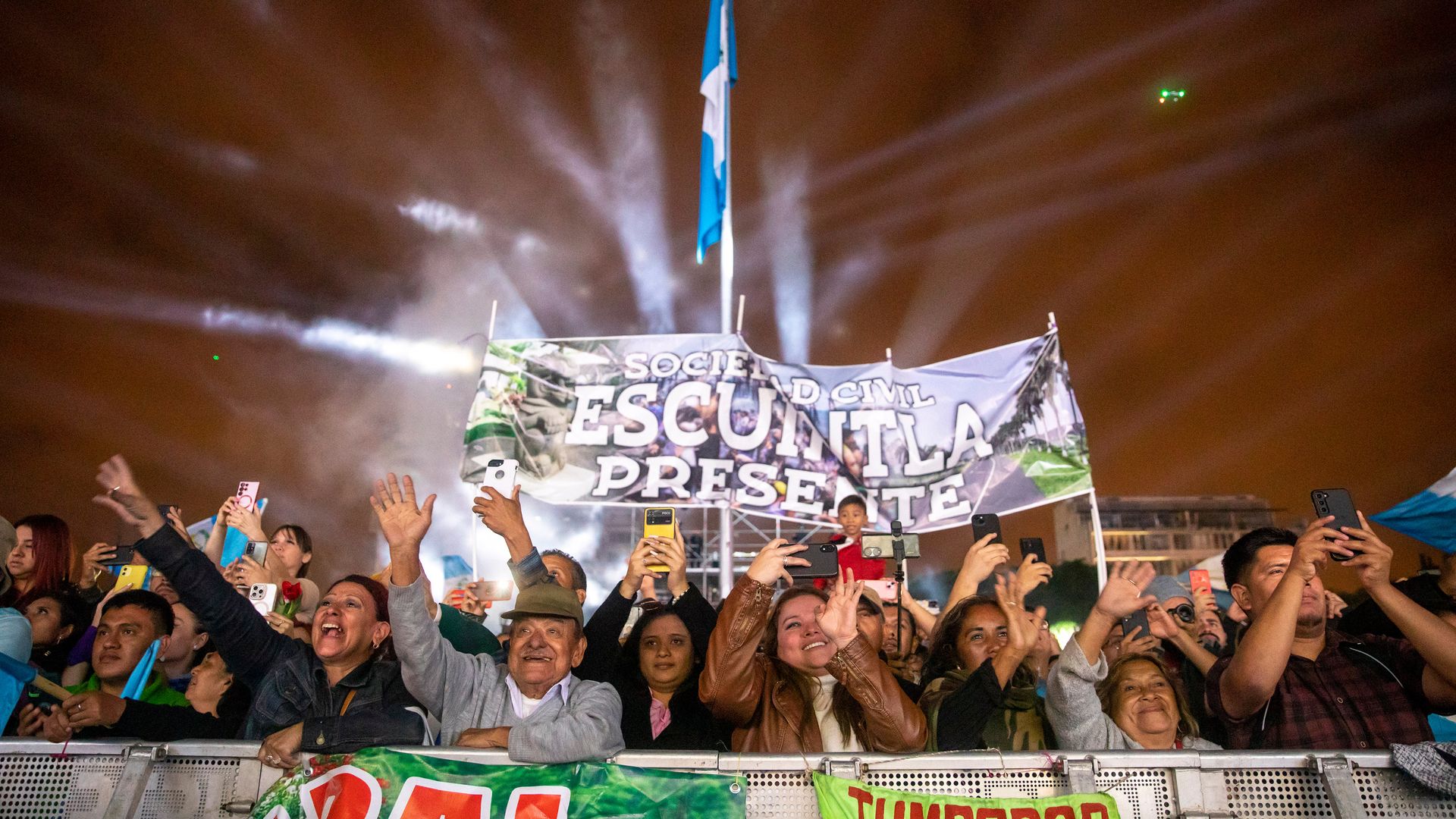 people who are lined up behind barriers raise their hands and cheer as President Bernardo Arévalo was inaugurated on Sunday. It is nighttime and fireworks are going off in the background.