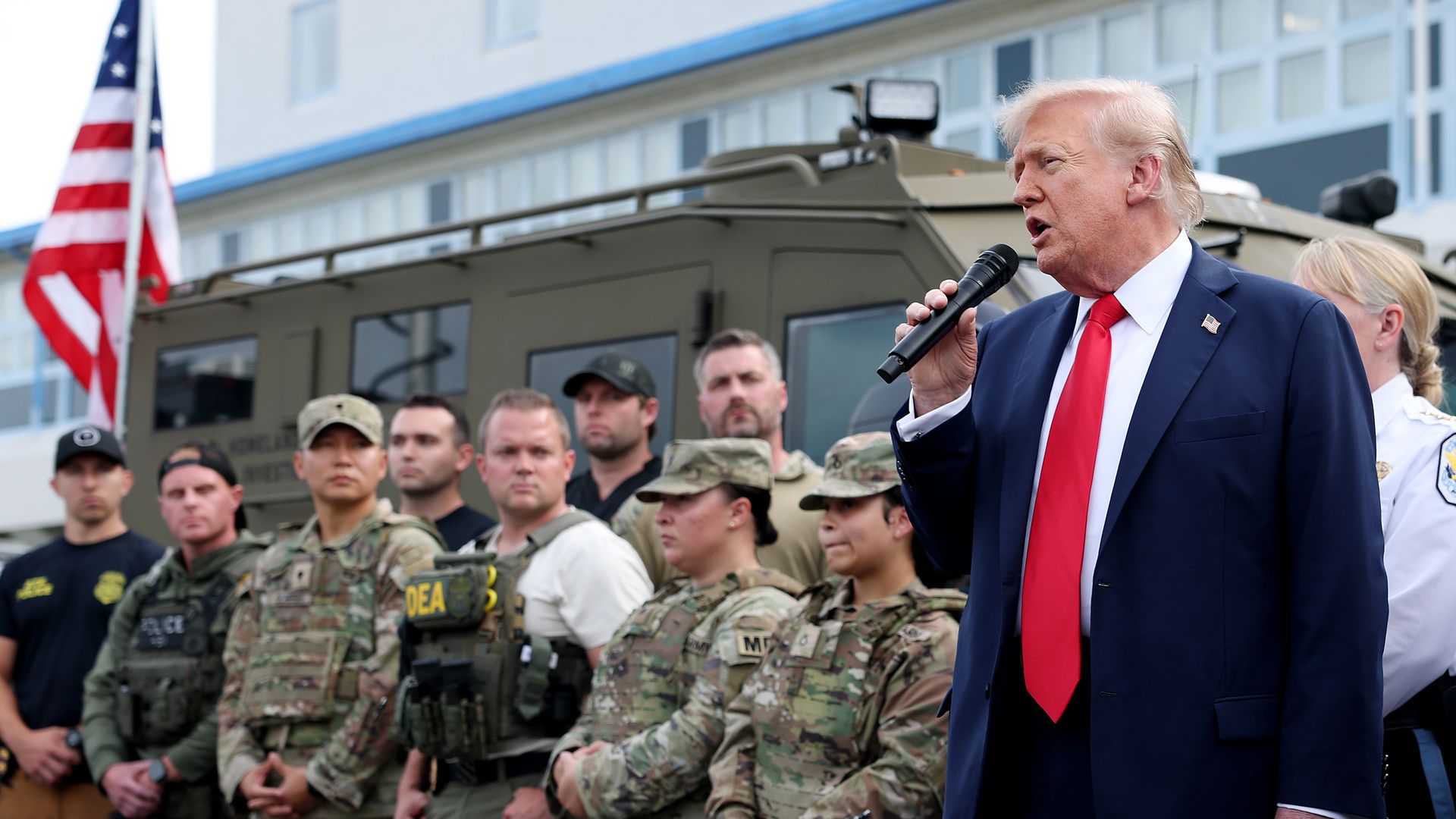 Man in navy suit and red tie speaking into microphone in front of a group of uniformed military and law enforcement personnel standing by a vehicle with an American flag nearby.