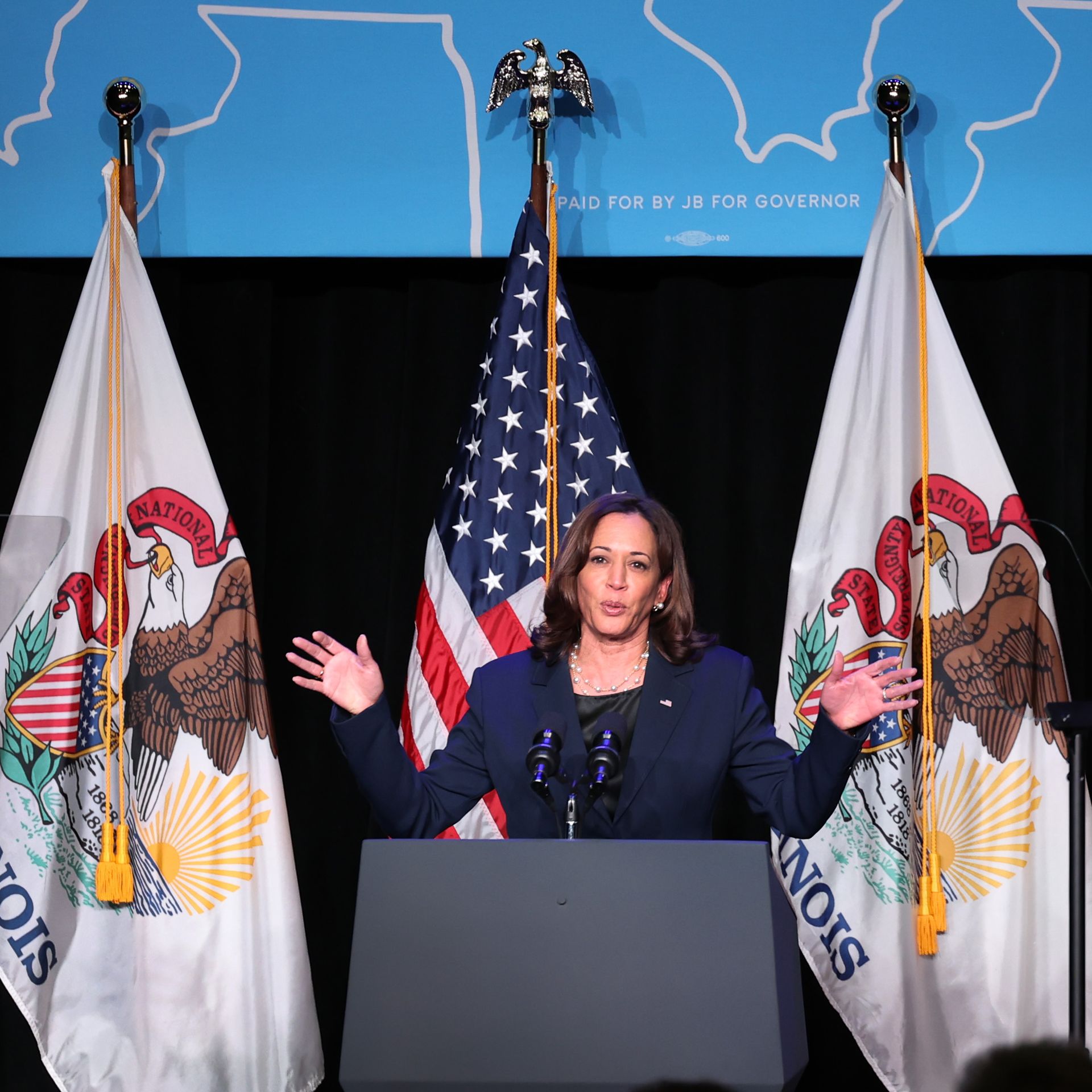 Vice President Harris in a navy blue suit standing behind a podium with the American flag behind her.