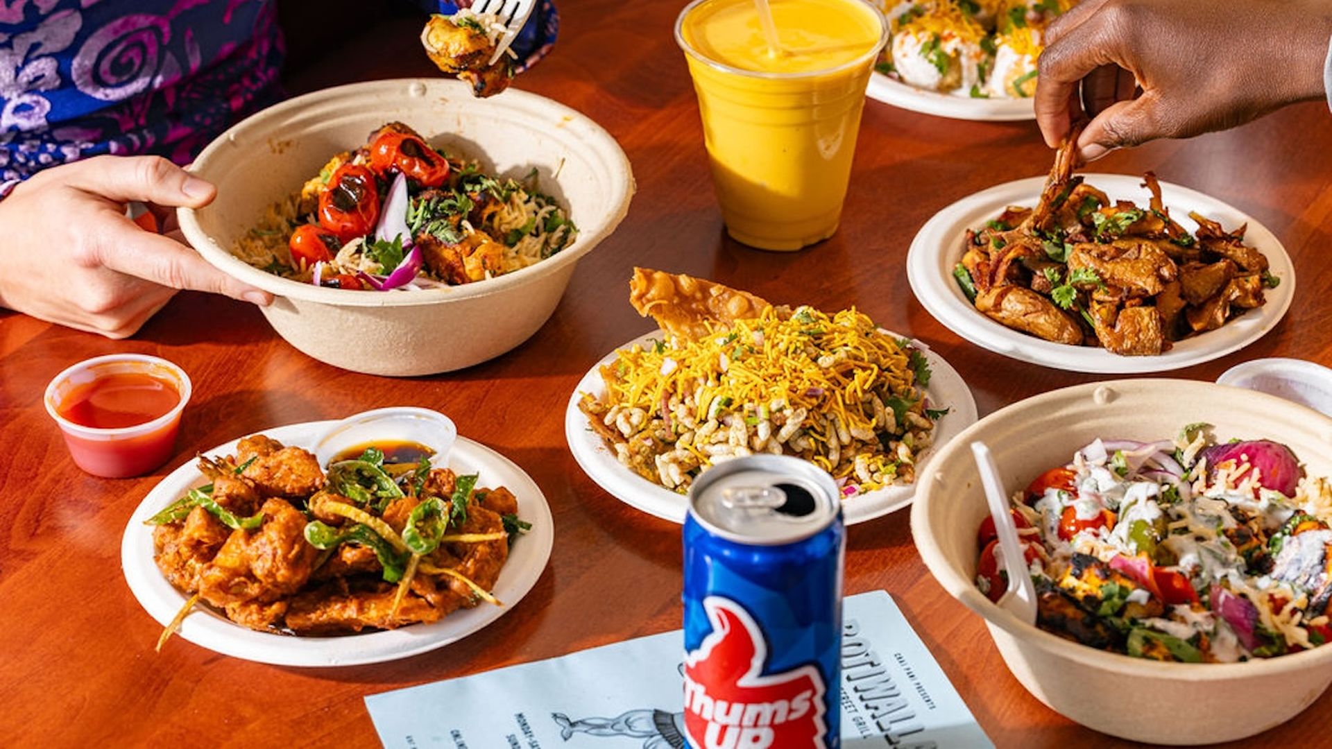 Table with assorted dishes: a large veggie rice bowl, crispy fried chicken, grilled meat plate, spicy salad bowl, cheesy fried rice, a yellow drink, and a blue Thums Up can; hands reach for food.