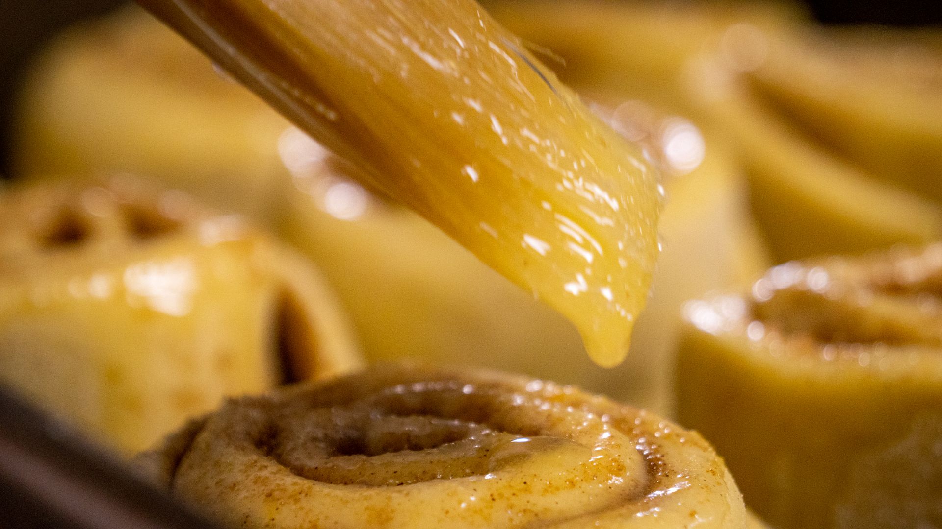 Close-up of a golden cinnamon roll being brushed with glossy glaze, with a softly blurred warm pastry background.
