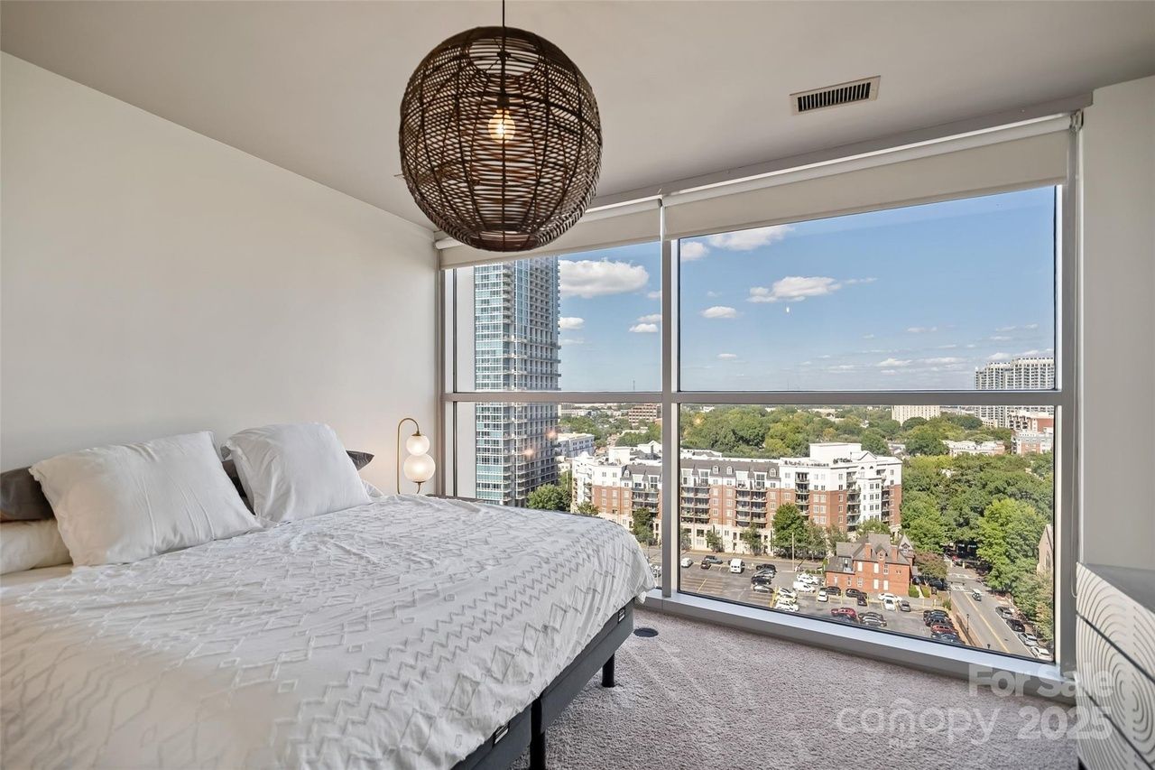 Modern bedroom with large window offering cityscape view, white bedding, beige carpet, and a round woven pendant light hanging from ceiling.