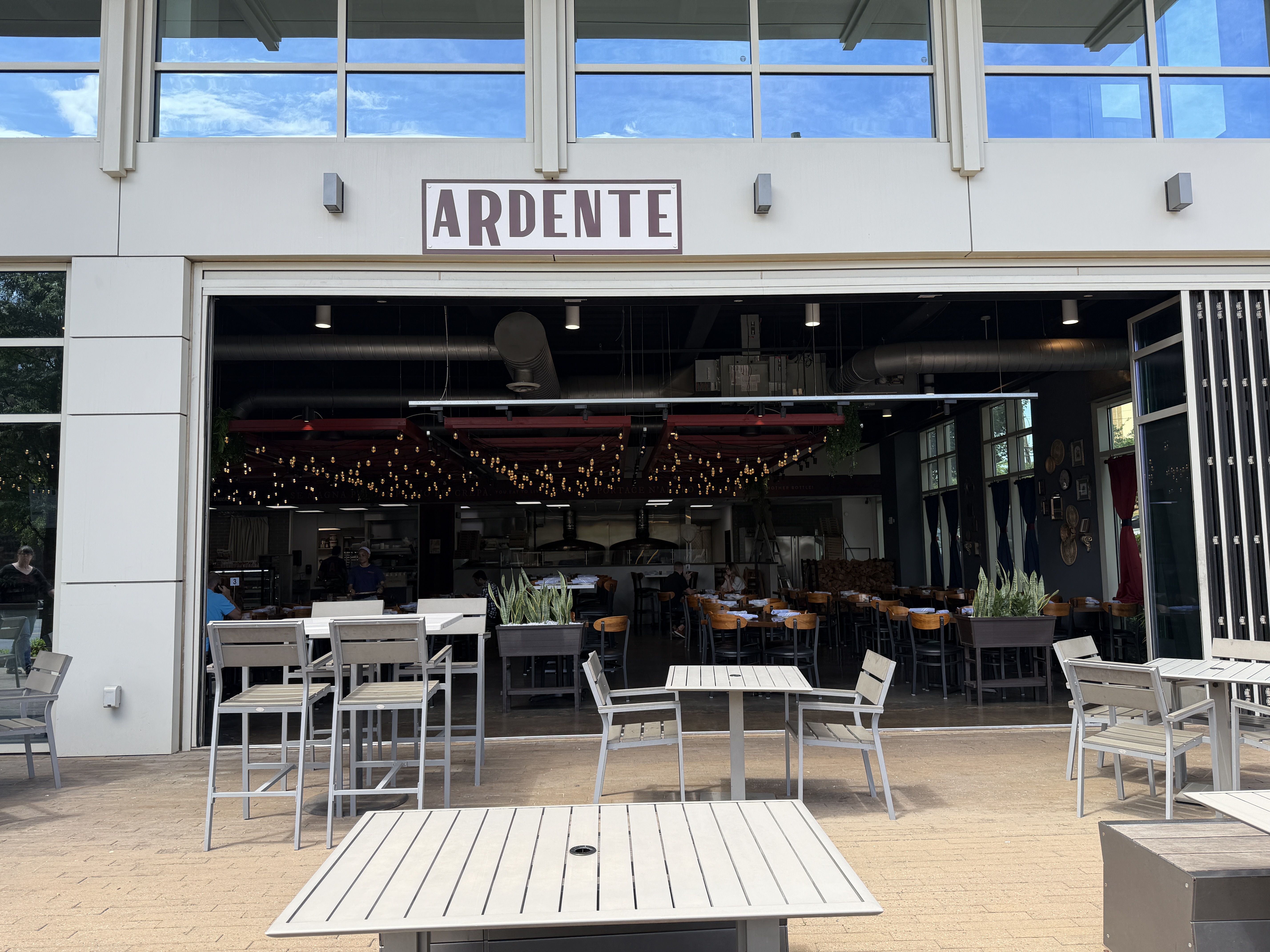 Front view of a modern cafe with an open storefront and a large sign reading "ARDENTE" above the entrance. Outdoor metal tables and chairs; string lights inside; blue sky reflected in the windows.