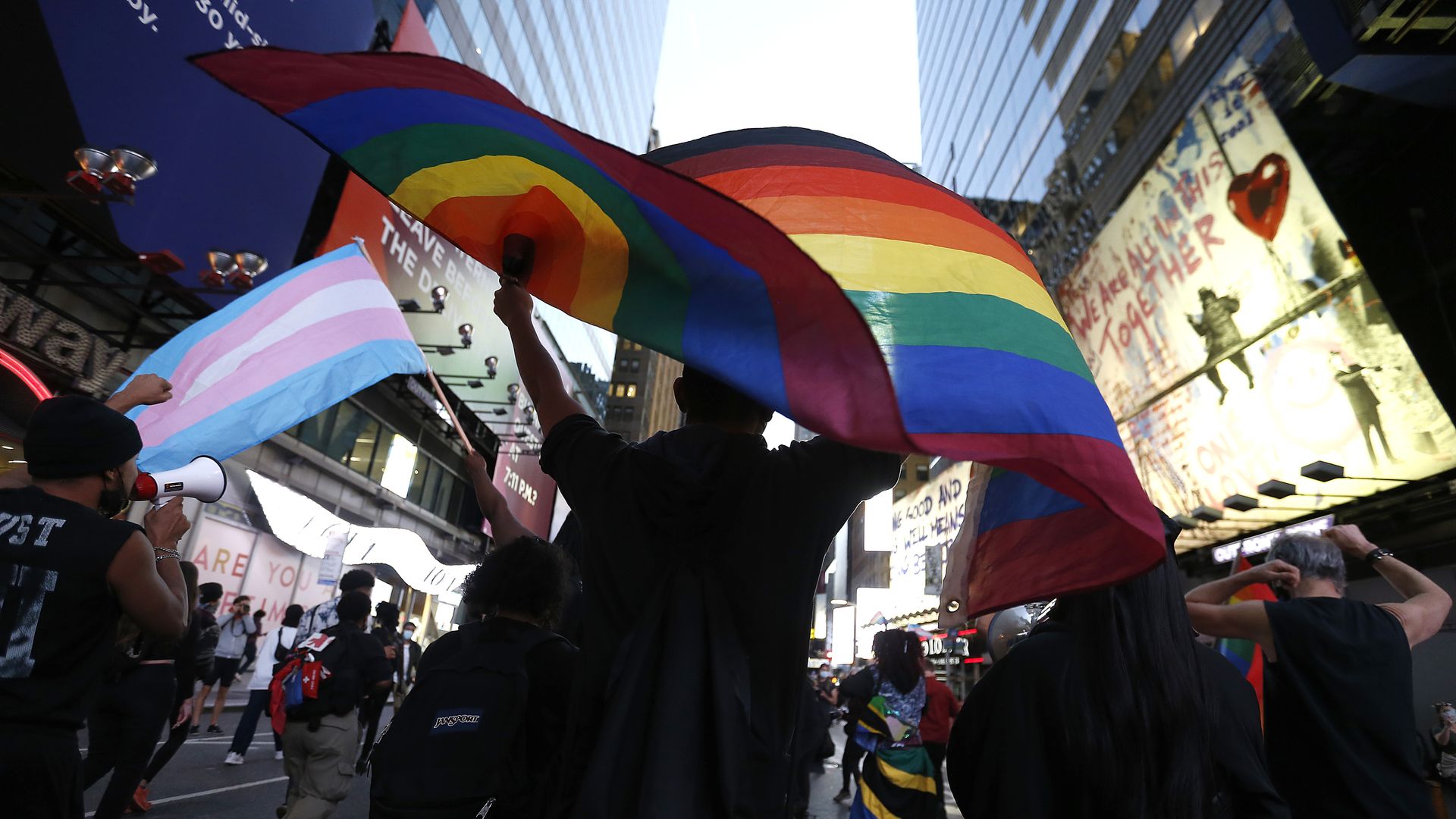 Photo of a person holding a rainbow flag in the air among a group of protesters. The trans flag is also visible.