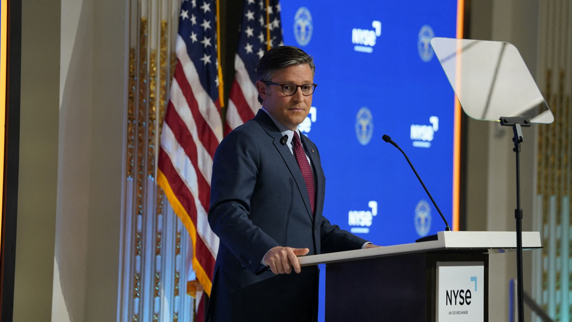 House Speaker Mike Johnson, wearing a blue suit, standing at an NYSE podium behind a glass teleprompter and in front of a blue backdrop, American flags and a white and gold wall. 