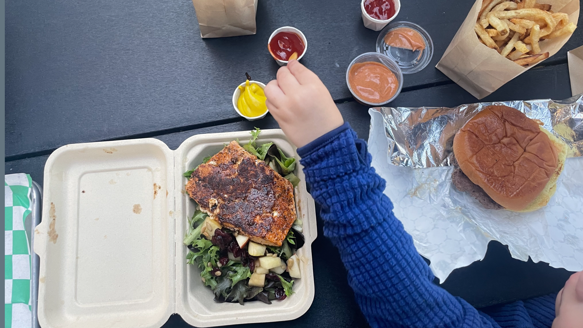 Child hand dipping a fry in ketchup from above with a salad with salmon and a burger