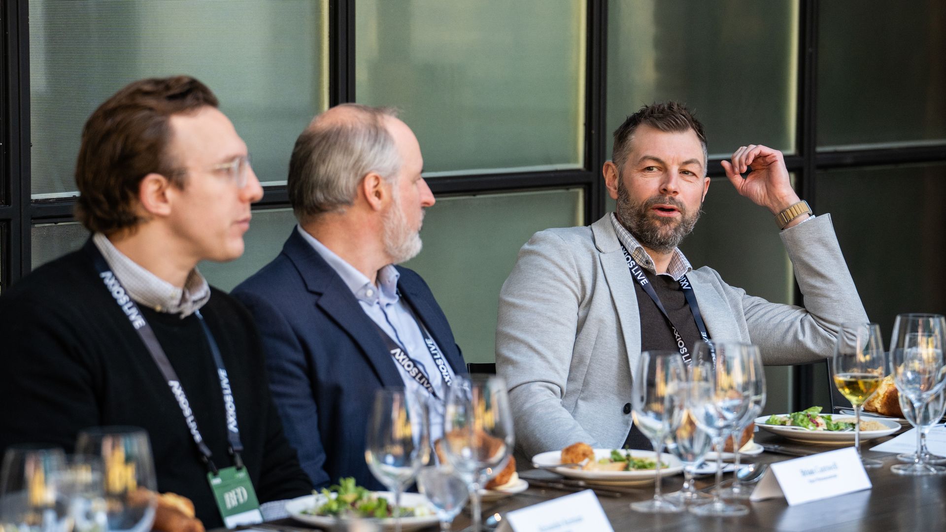 Three men sit at a table engaged in conversation, near a windowed wall at a professional luncheon.