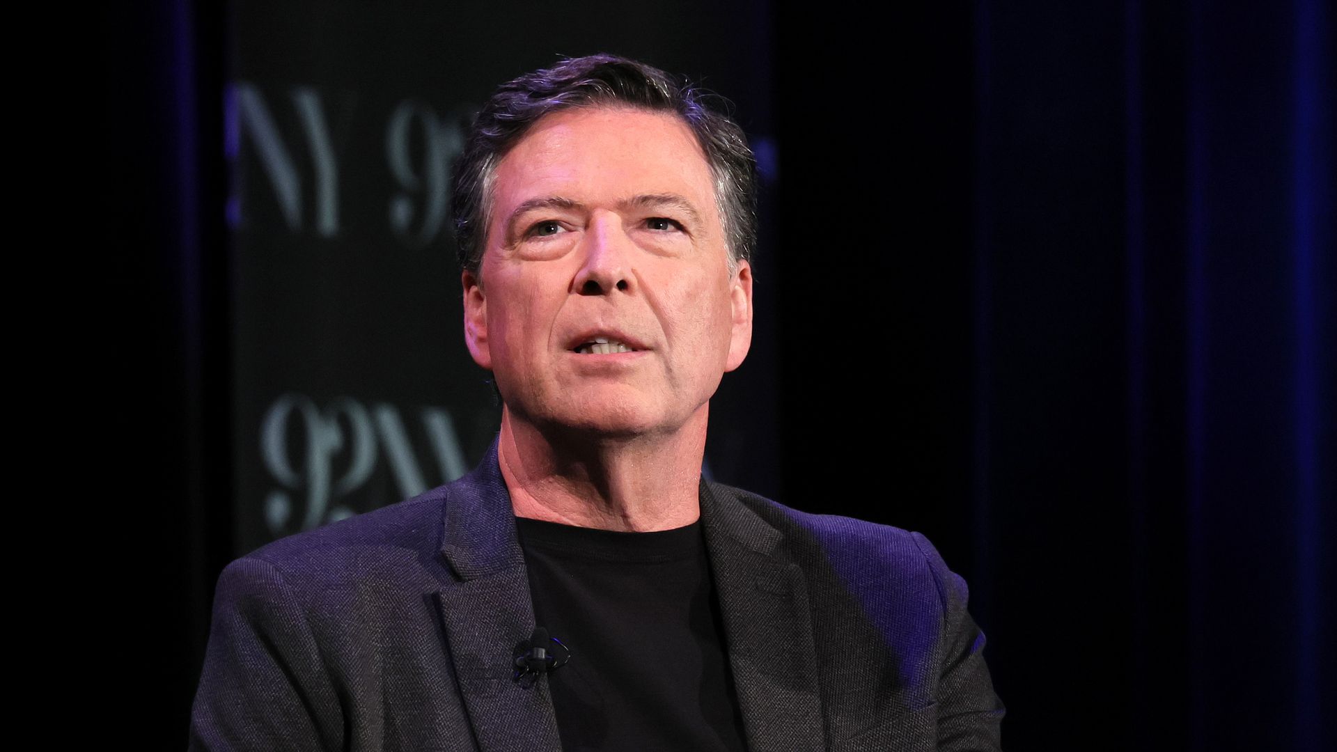 Former FBI Director James Comey, with dark, graying hair and wearing a black suit and black t-shirt, clasps his hands while speaking at an event.