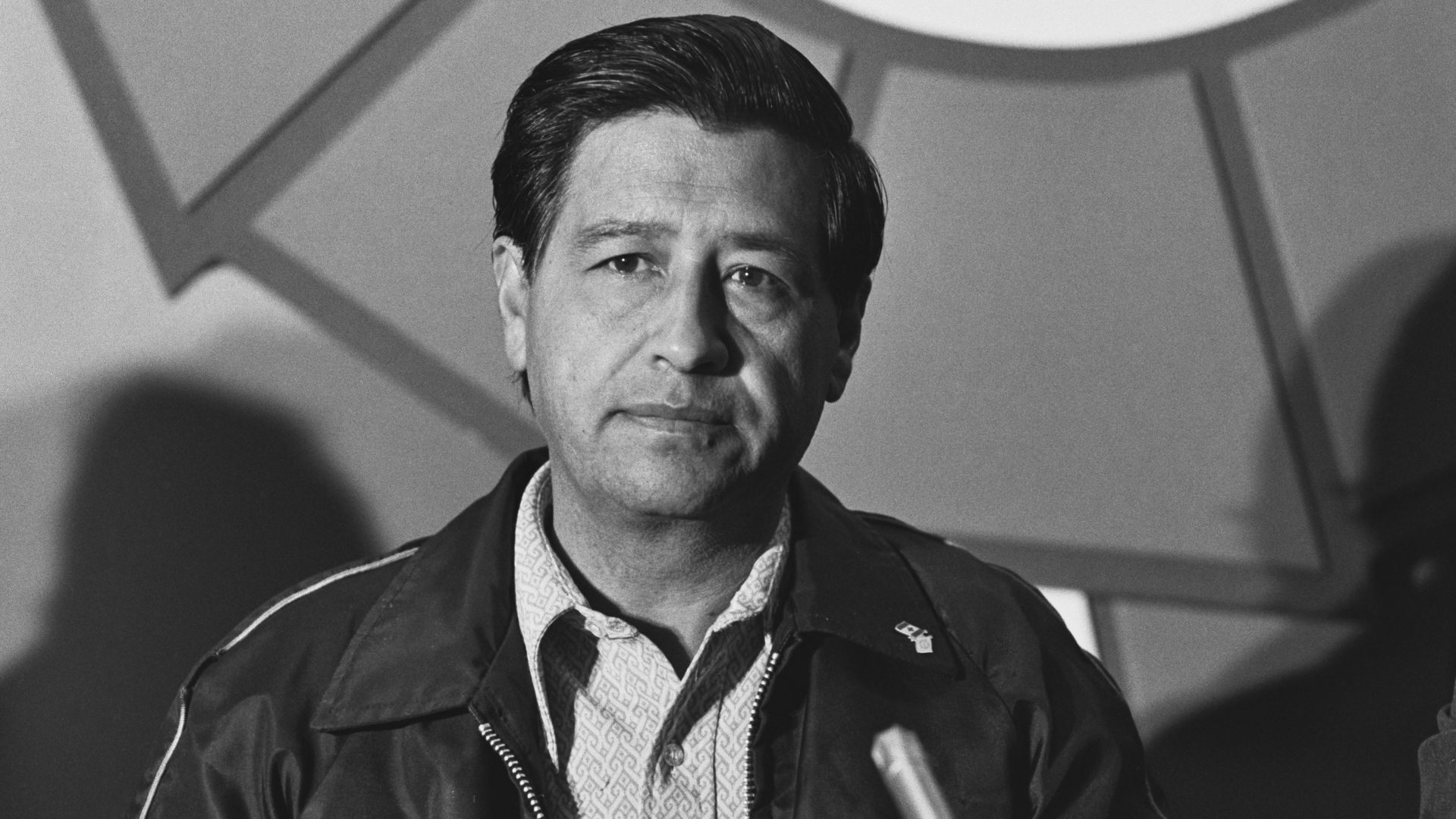 Black-and-white portrait of a middle-aged man with dark hair, wearing a patterned shirt and jacket, looking at the camera. Abstract geometric backdrop with intersecting shapes.