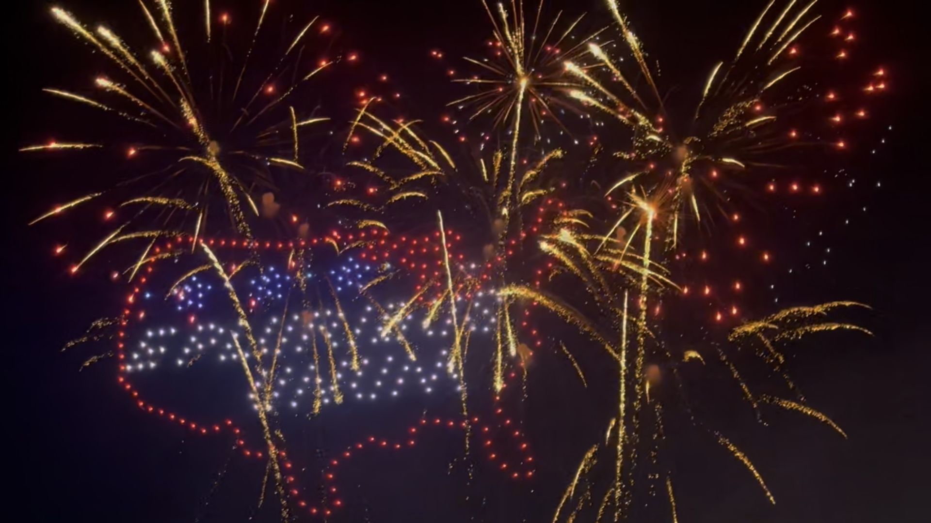 Fireworks explode across a dark night sky, with bright golden trails, red sparkles, and blue dots forming a festive display.