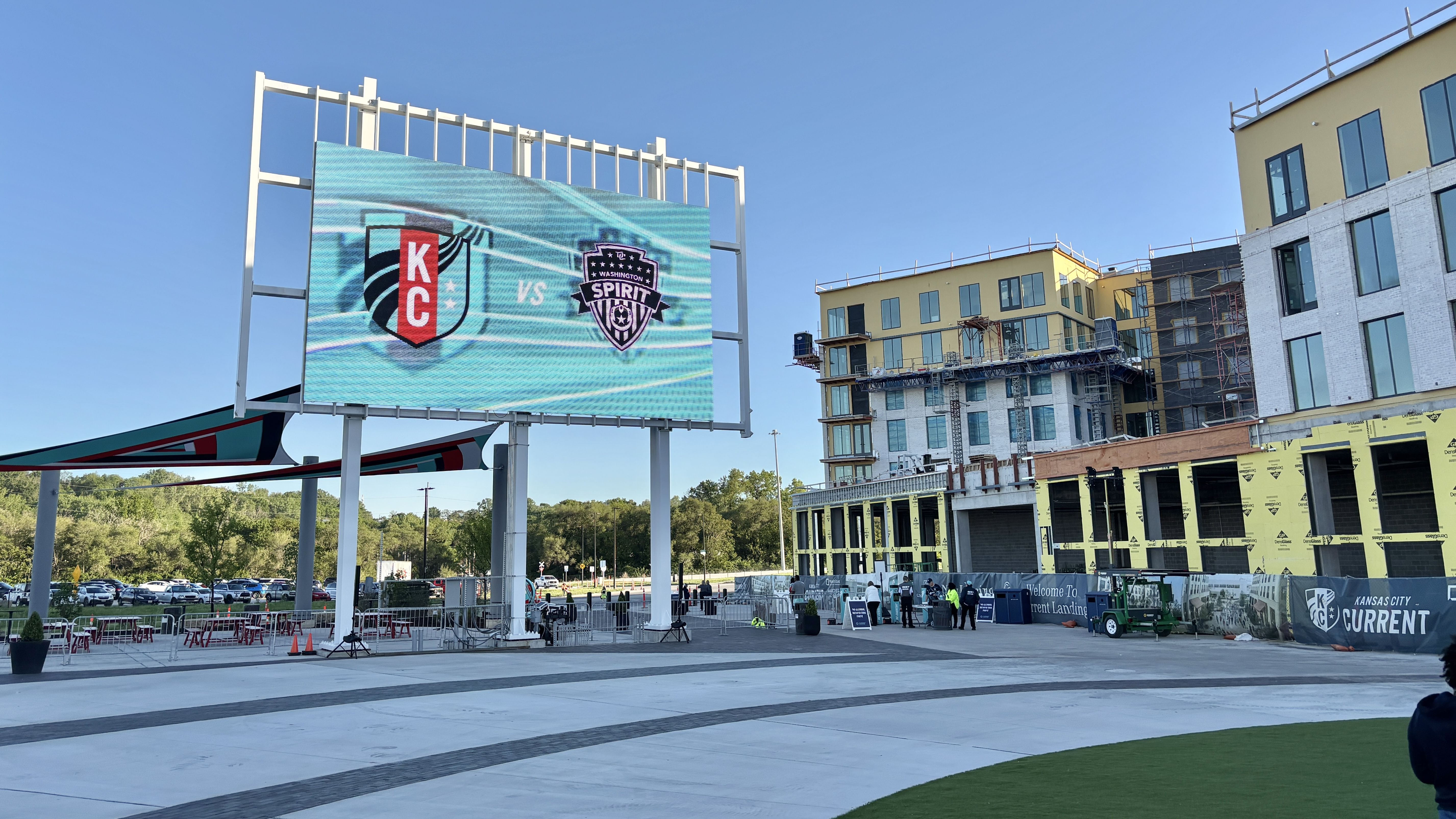 Outdoor plaza with a large LED screen displaying KC vs Spirit logos against a blue sky. Right side shows a building under construction with scaffolding; barriers and people near the entrance.
