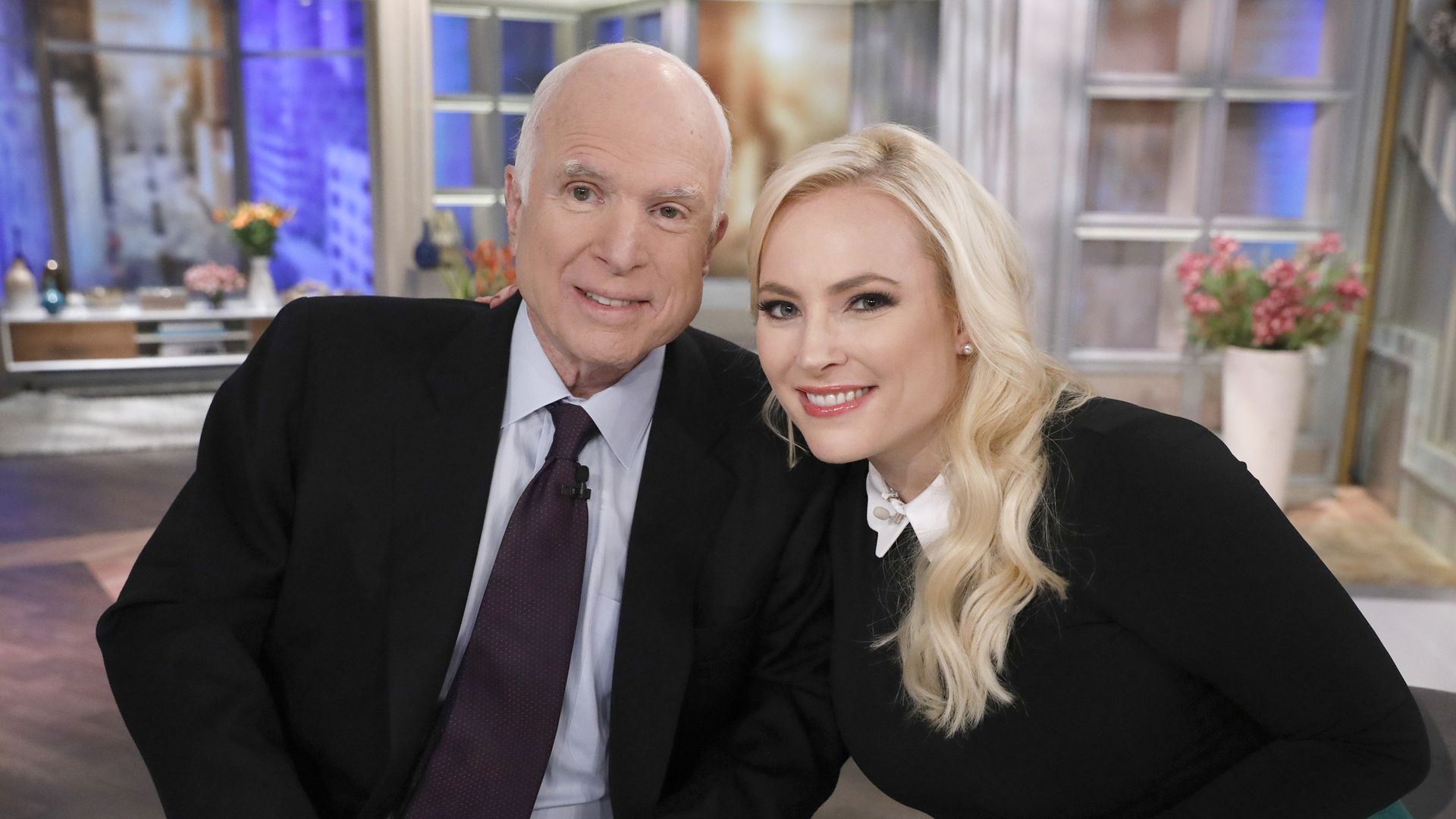 An older man in a dark suit and a blonde woman in a black top with a floral skirt smile at the camera while sitting at a glass table with blue mugs, a vase of flowers behind them.