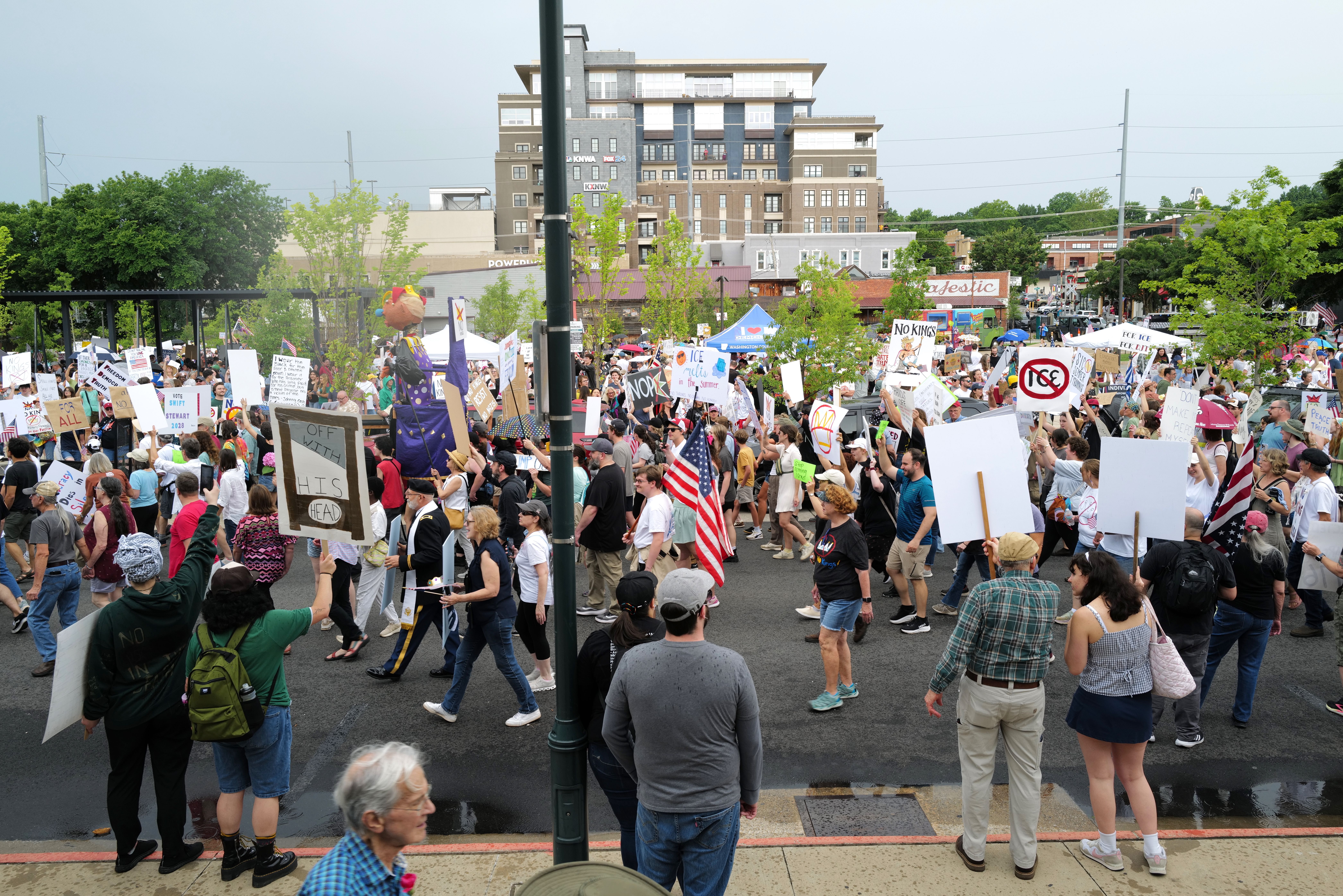 Large crowd marches with protest signs, American flags, and a king puppet, calling for democracy and rejecting tyranny.