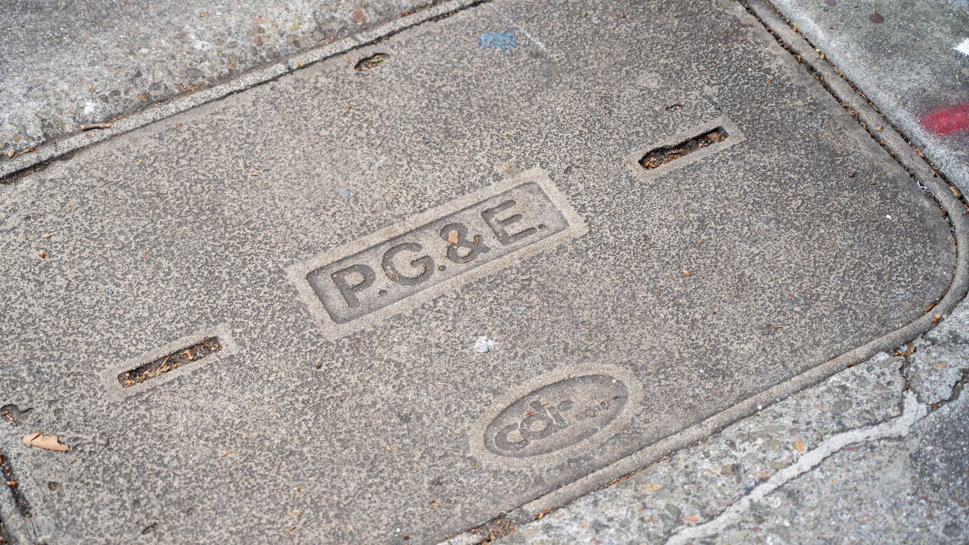Weathered gray utility cover on a sidewalk, with PG&E embossed in a rectangle and a circular logo nearby; dirt, leaves, and cracks around.
