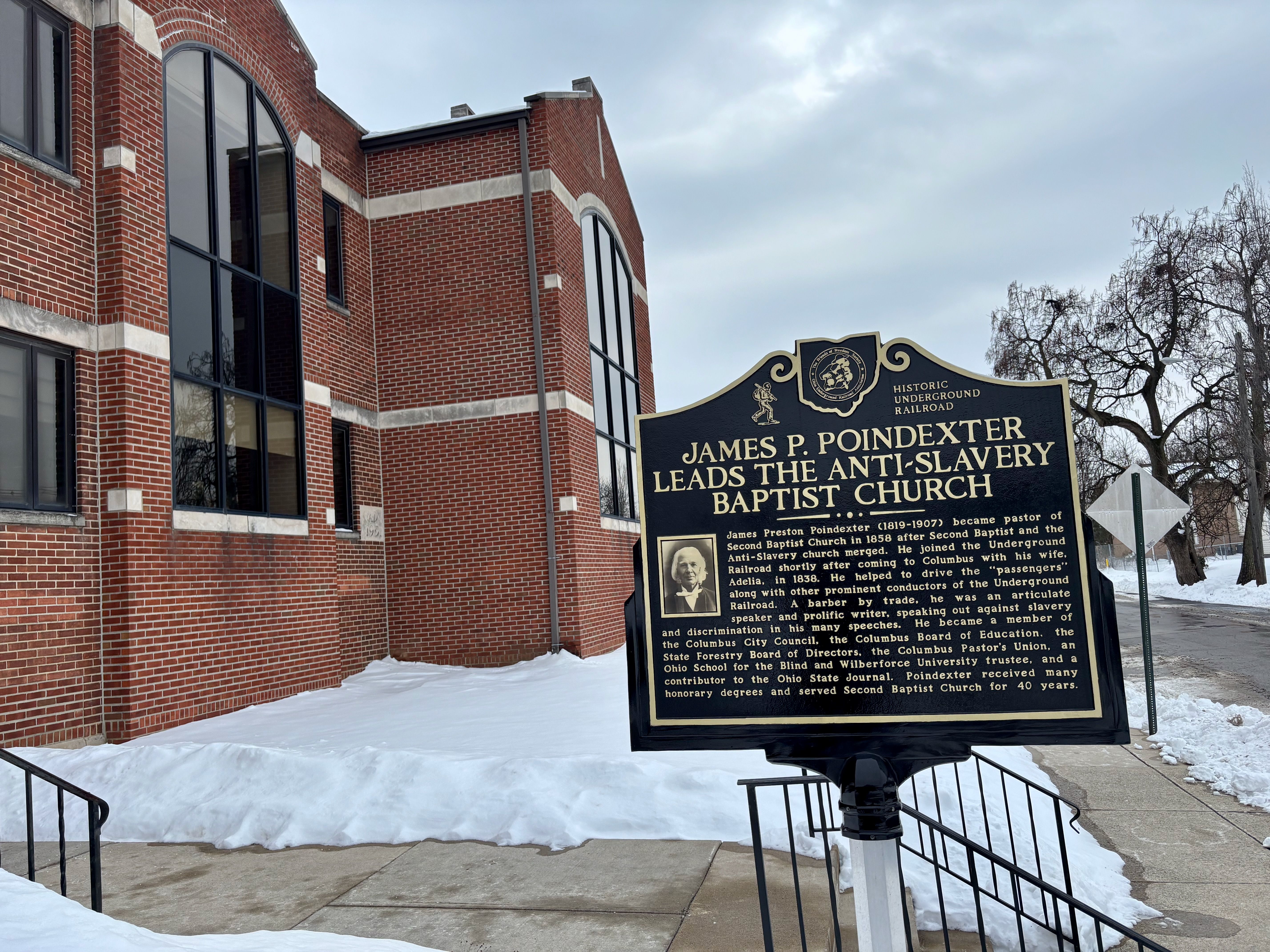 Black historical marker about James P. Poindexter leading the anti-slavery Baptist Church near red brick building, snow on ground, overcast sky, sidewalk and leafless trees nearby.
