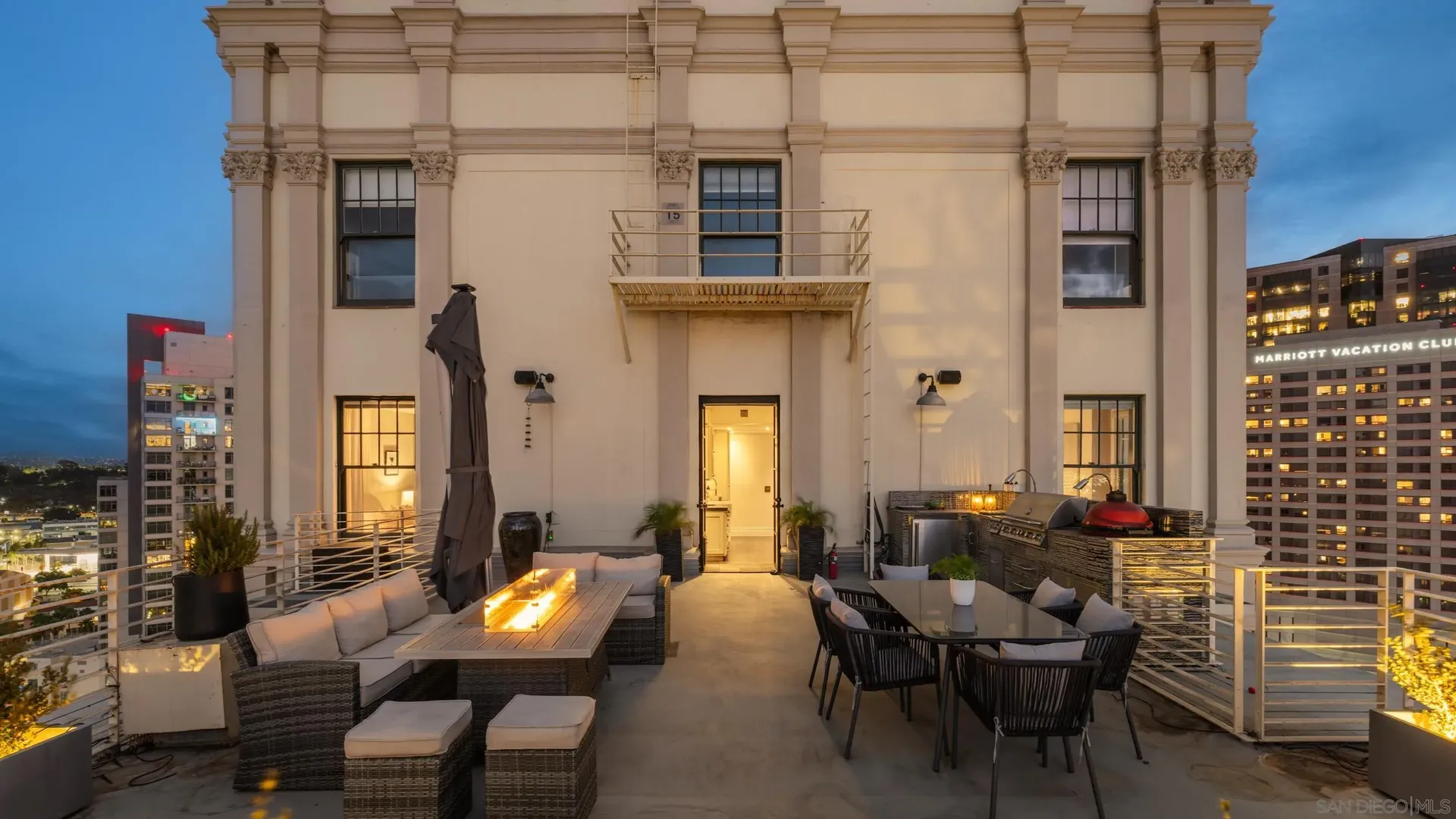 Rooftop terrace on a cream ornate building with decorative crown molding; a blue evening sky. Wicker sofas and chairs surround a glowing fire table, with a grill on the right and distant city buildings.