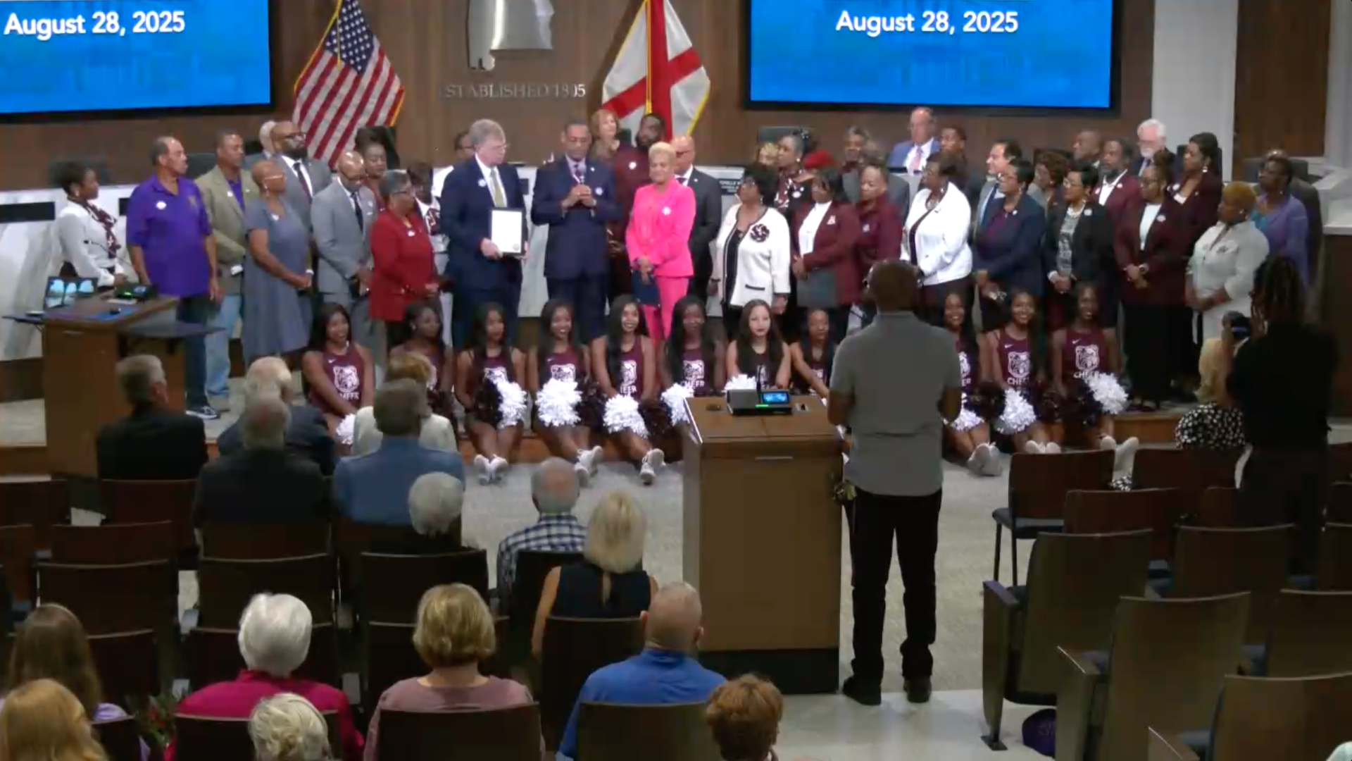 Group photo at an event with cheerleaders in maroon uniforms seated front, a diverse group of adults standing behind them, two screens display August 28, 2025, with American and Alabama flags present.