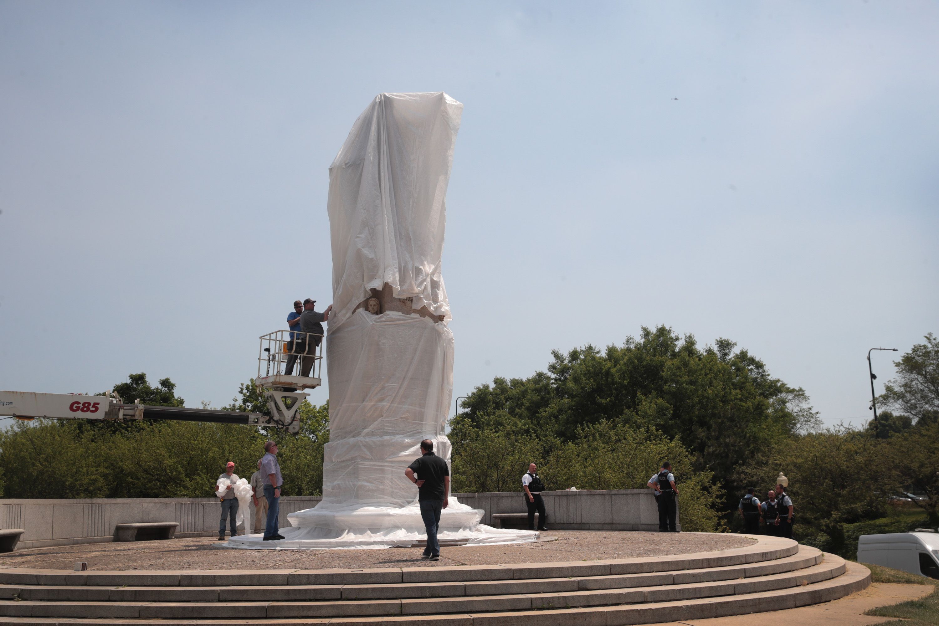 Workers on a lift and ground cover a tall statue with white plastic wrap on a stepped circular platform, with police and bystanders nearby and trees in the background under a clear sky.