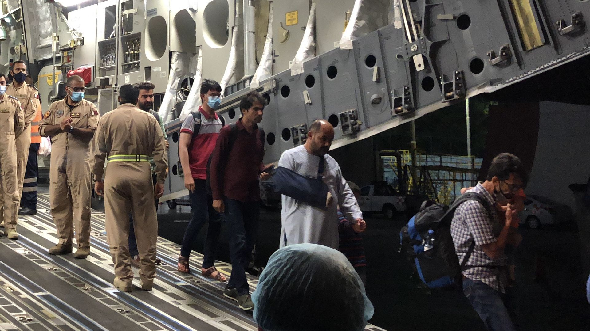 Group of people in beige uniforms and face masks walk beside a large gray military aircraft, its interior exposed with equipment, ladders, and circular cutouts along the fuselage.
