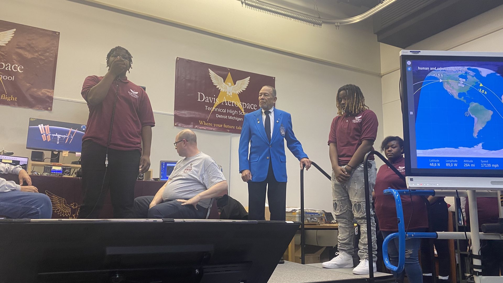 A Davis student is shown speaking into a microphone of a ham radio while another student waits. Radio operators are in the background. Everyone is on a stage. 