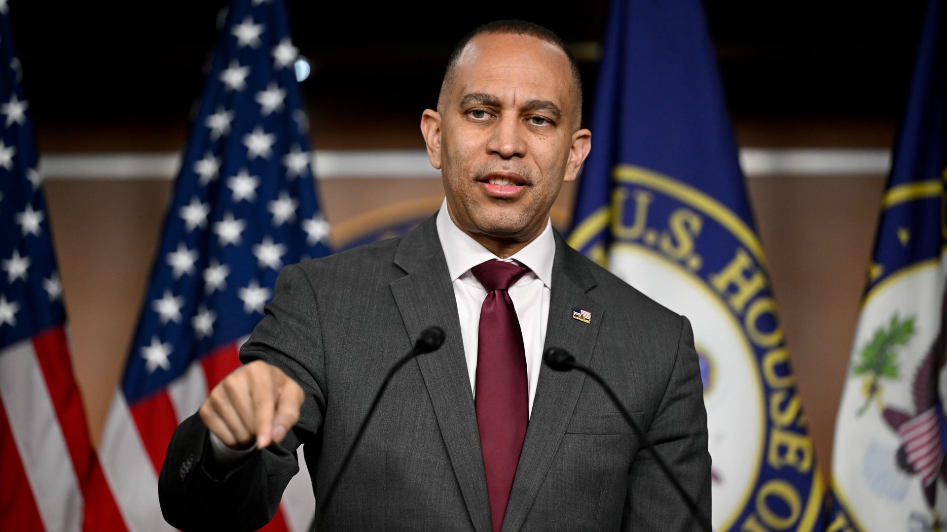 A man in a gray suit and burgundy tie speaks at a podium with two microphones, gesturing with his right hand. Behind him are American flags and a U.S. House seal.
