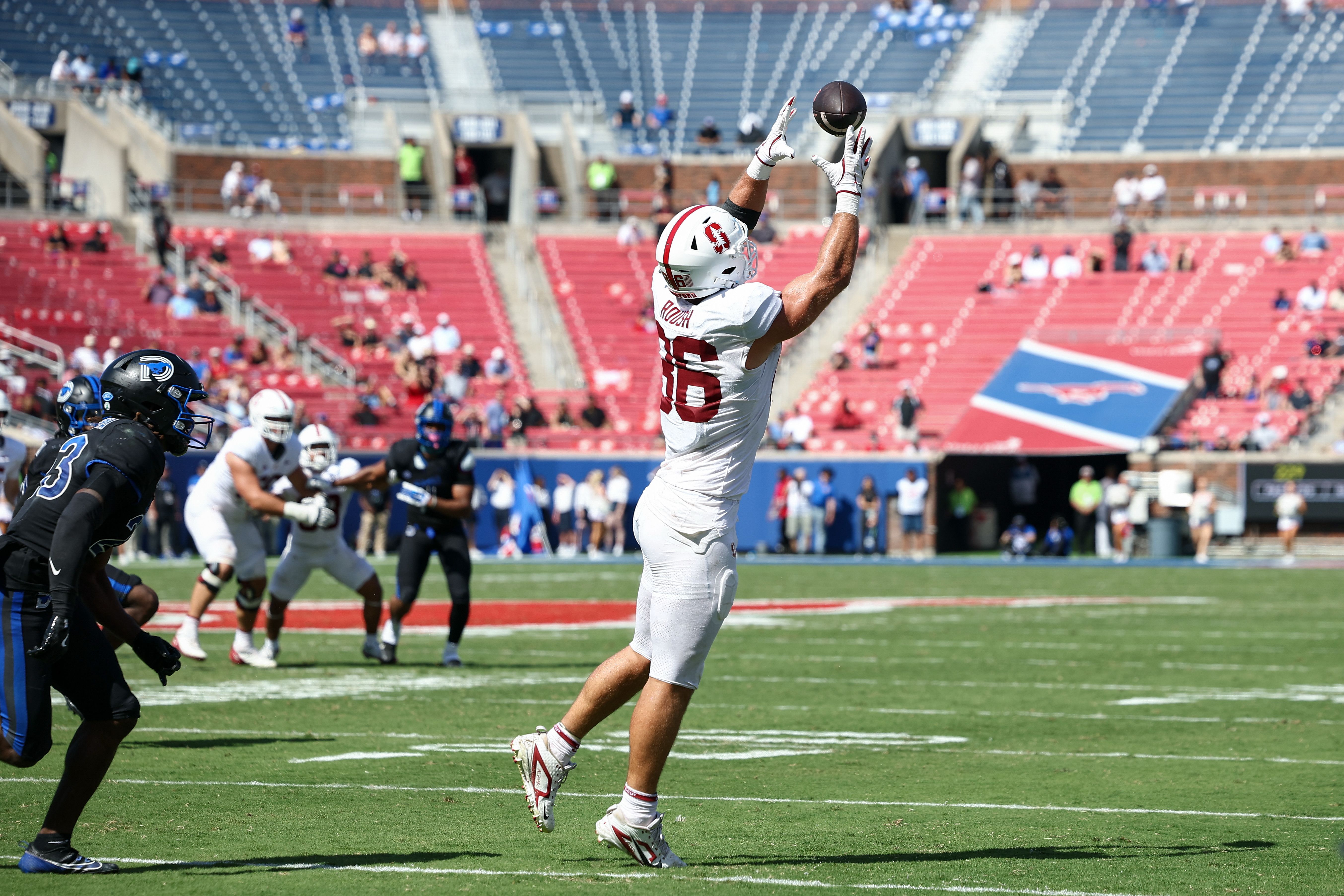A football player in a white uniform leaps to catch a ball above the crowd, arms outstretched; black-clad defenders near, red stadium seats and fans in the background.