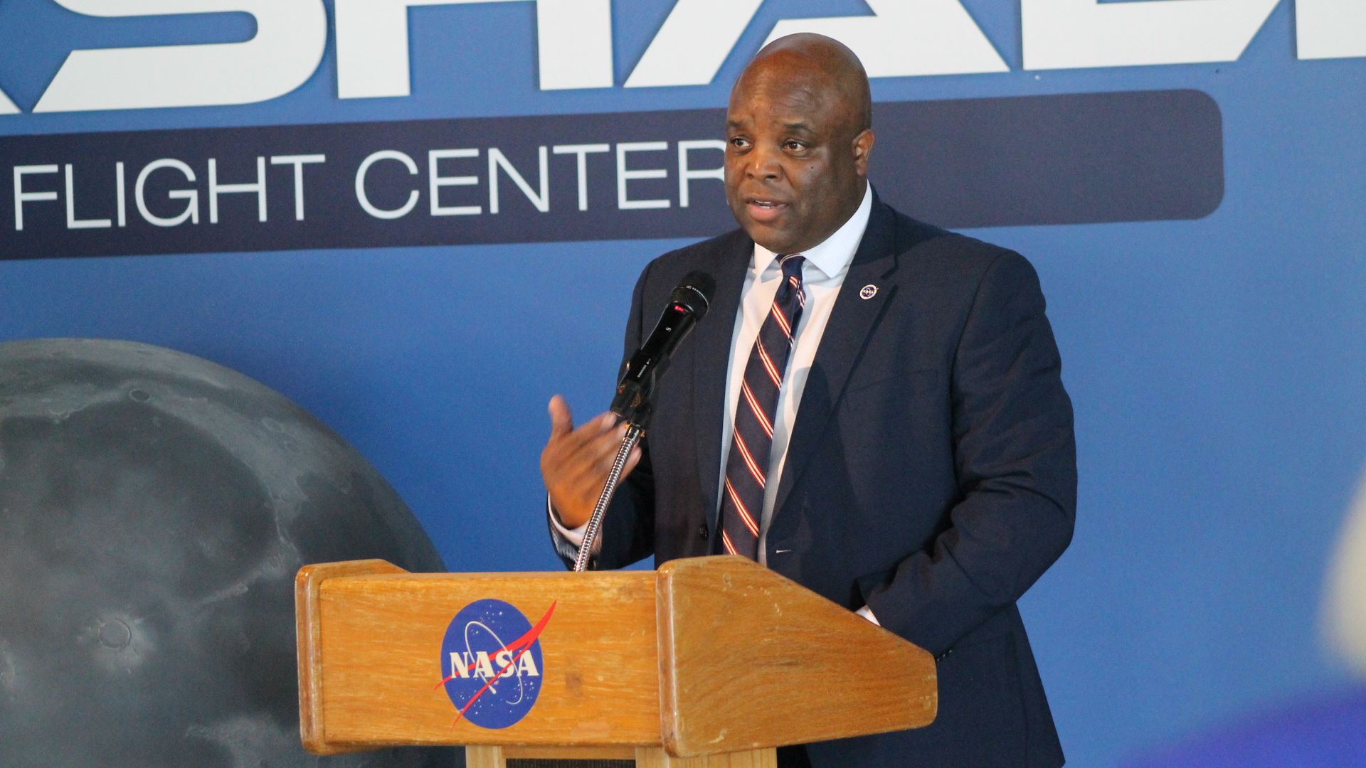 Man in dark suit and striped tie speaking at a NASA wooden podium with microphone, blue background with large white text and image of the moon