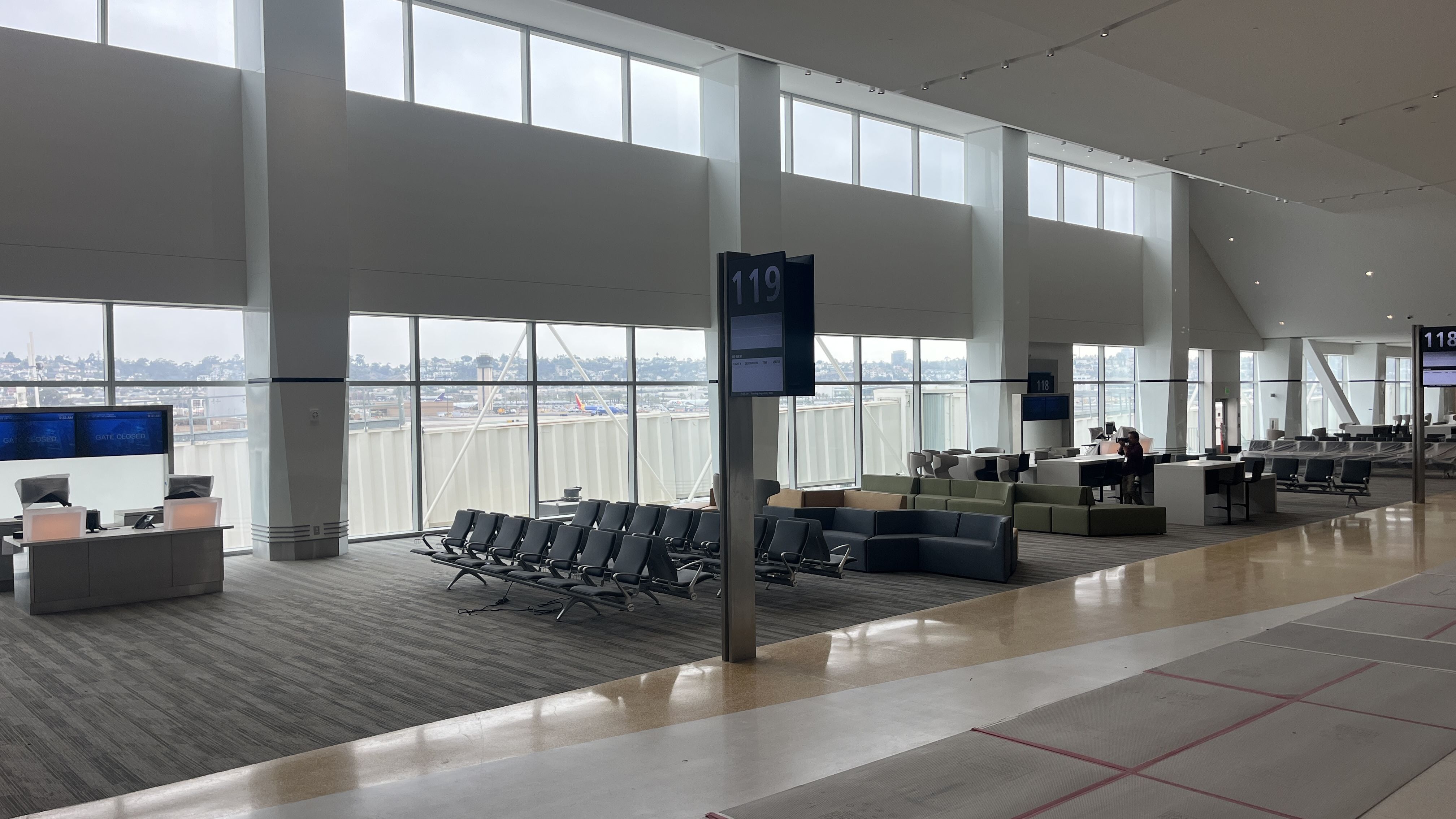 Empty airport gate area with rows of black seats, large windows showing a cloudy sky and cityscape, gate signs numbered 118 and 119, and some tables and couches.