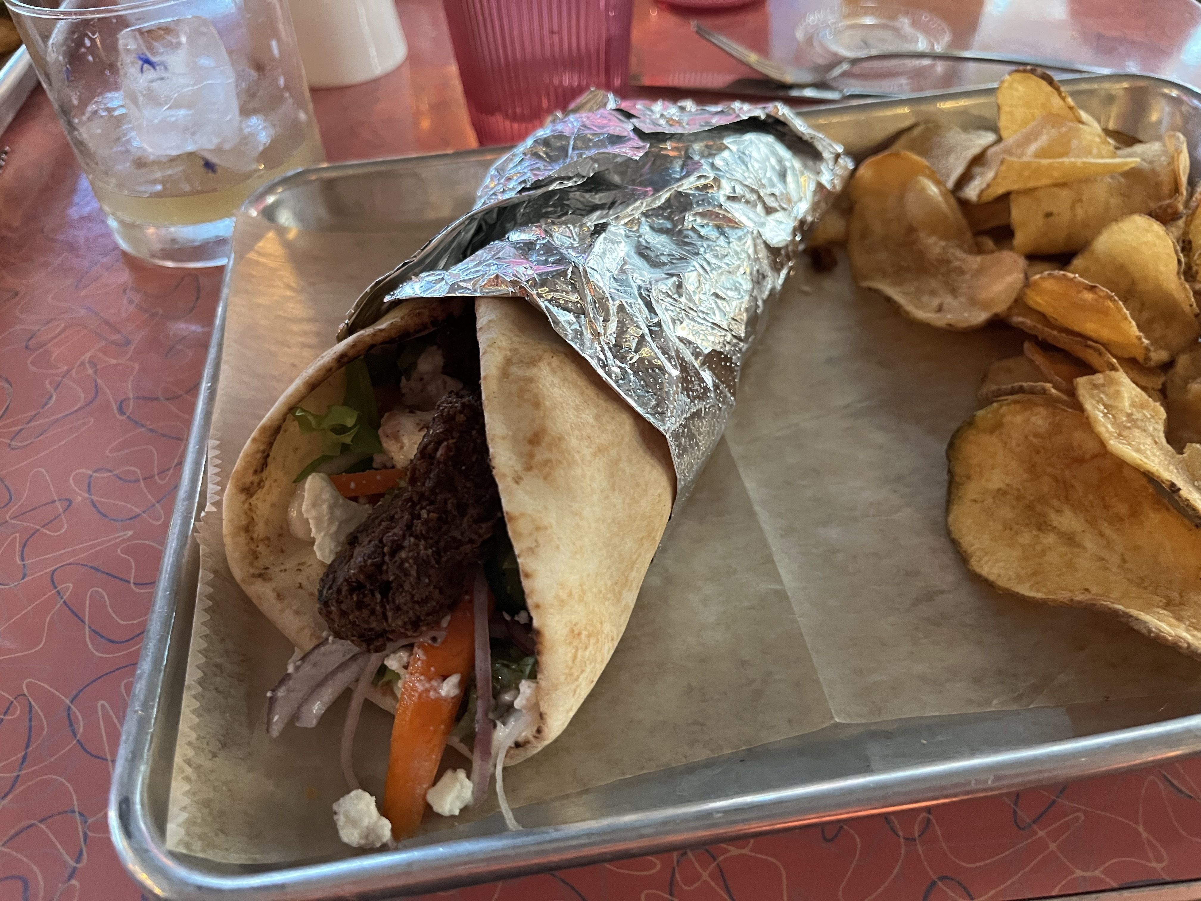 A falafal wrap in tinfoil and next to potato chips on a red table 