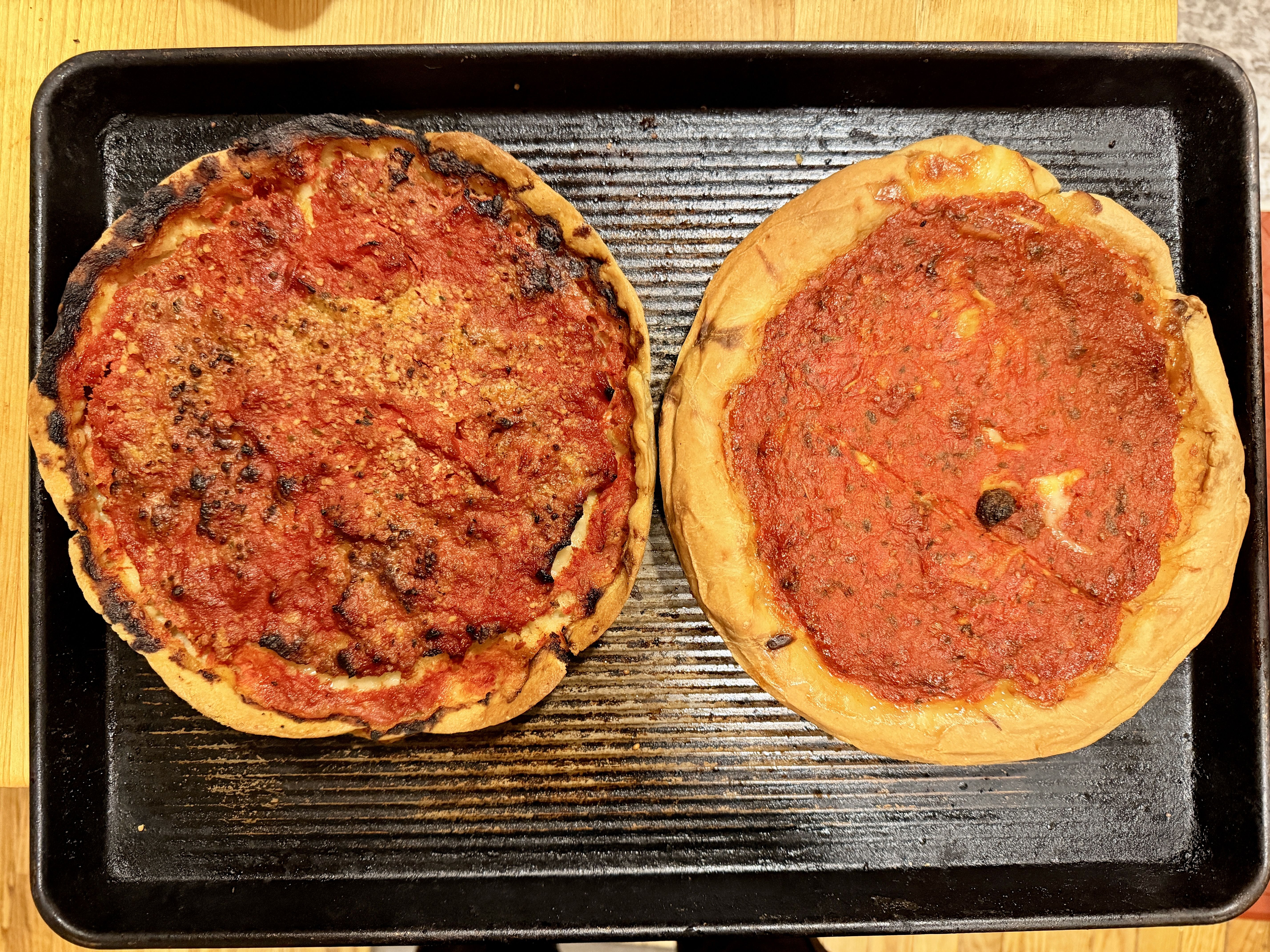 Two thick pizzas with red tomato sauce and melted cheese on a dark ridged grill pan, crusts golden with charred spots, set on a wooden table.