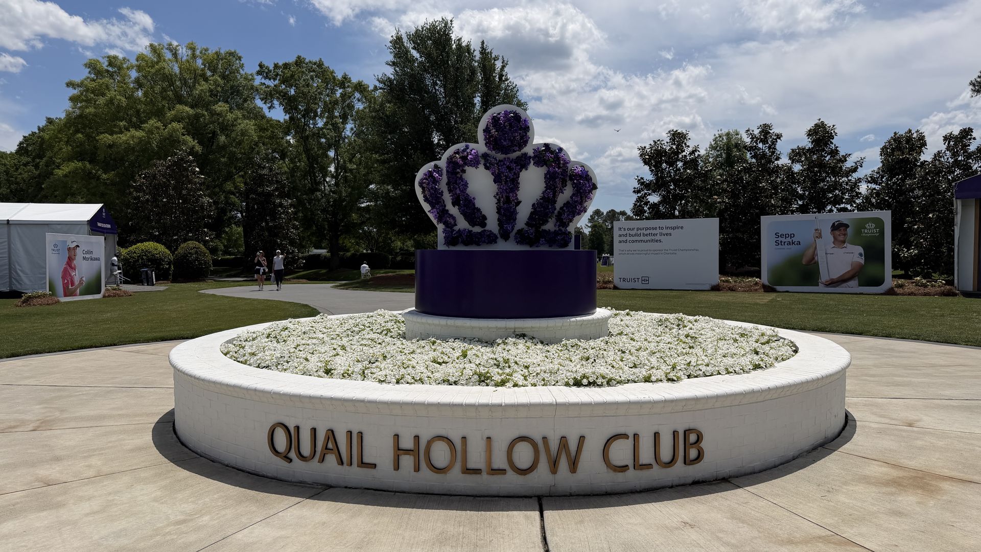 Round white planter with a purple floral crown sculpture at Quail Hollow Club; green trees, blue sky with clouds, and event banners in the background with a few people walking.