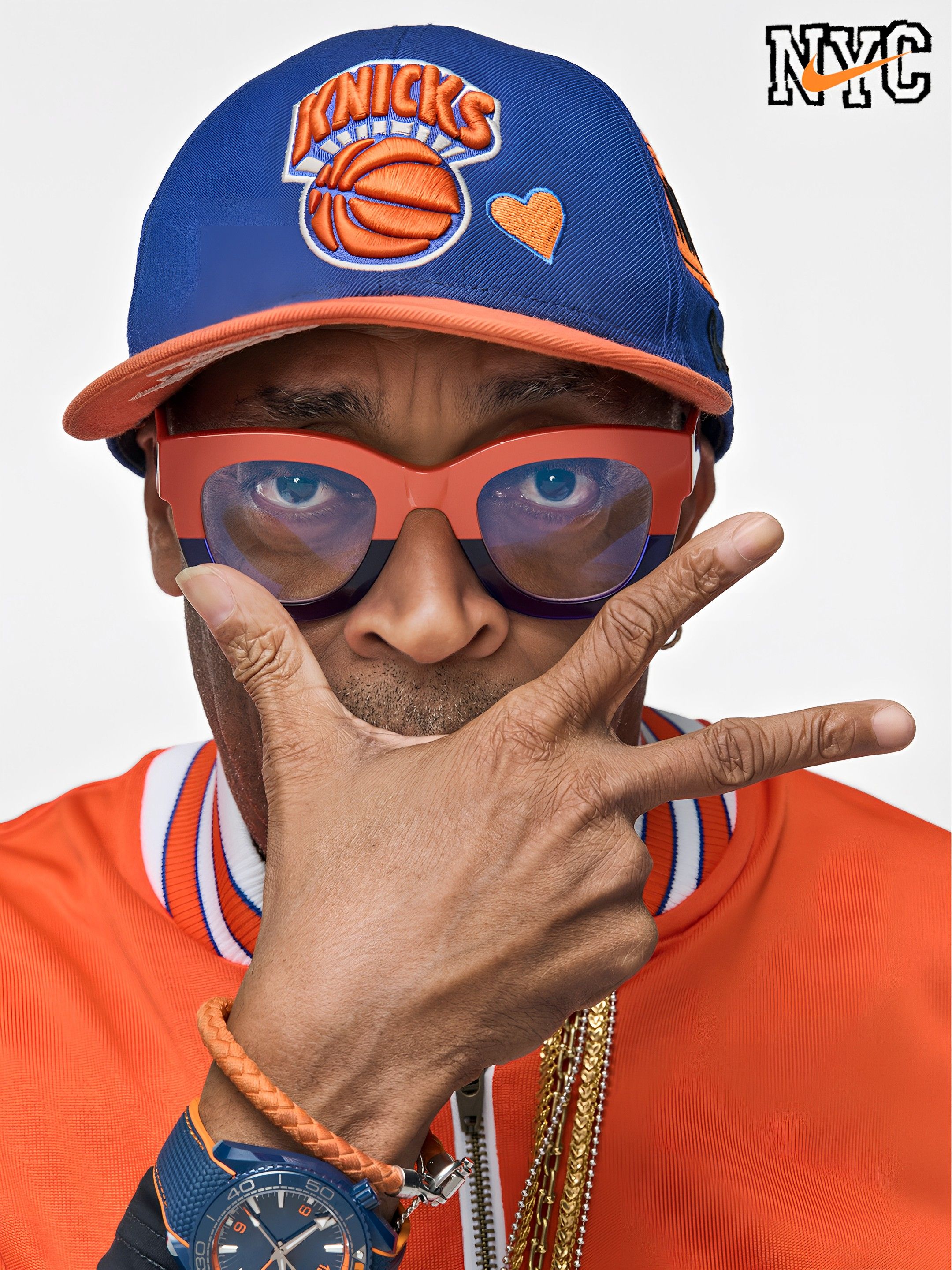 Close-up of a person wearing a blue Knicks cap with an orange basketball logo and orange heart patch, orange sunglasses, orange outfit with white-blue trim, and gold chains; NYC logo in corner.