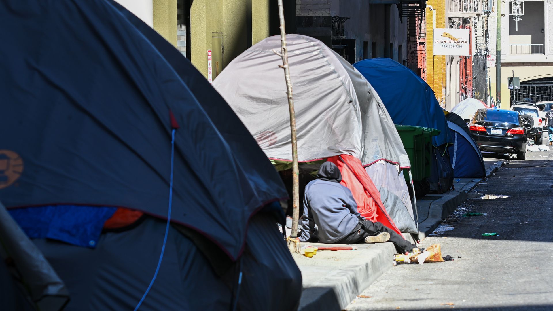Photo of a person sitting on a sidewalk outside a tent in a homeless encampment