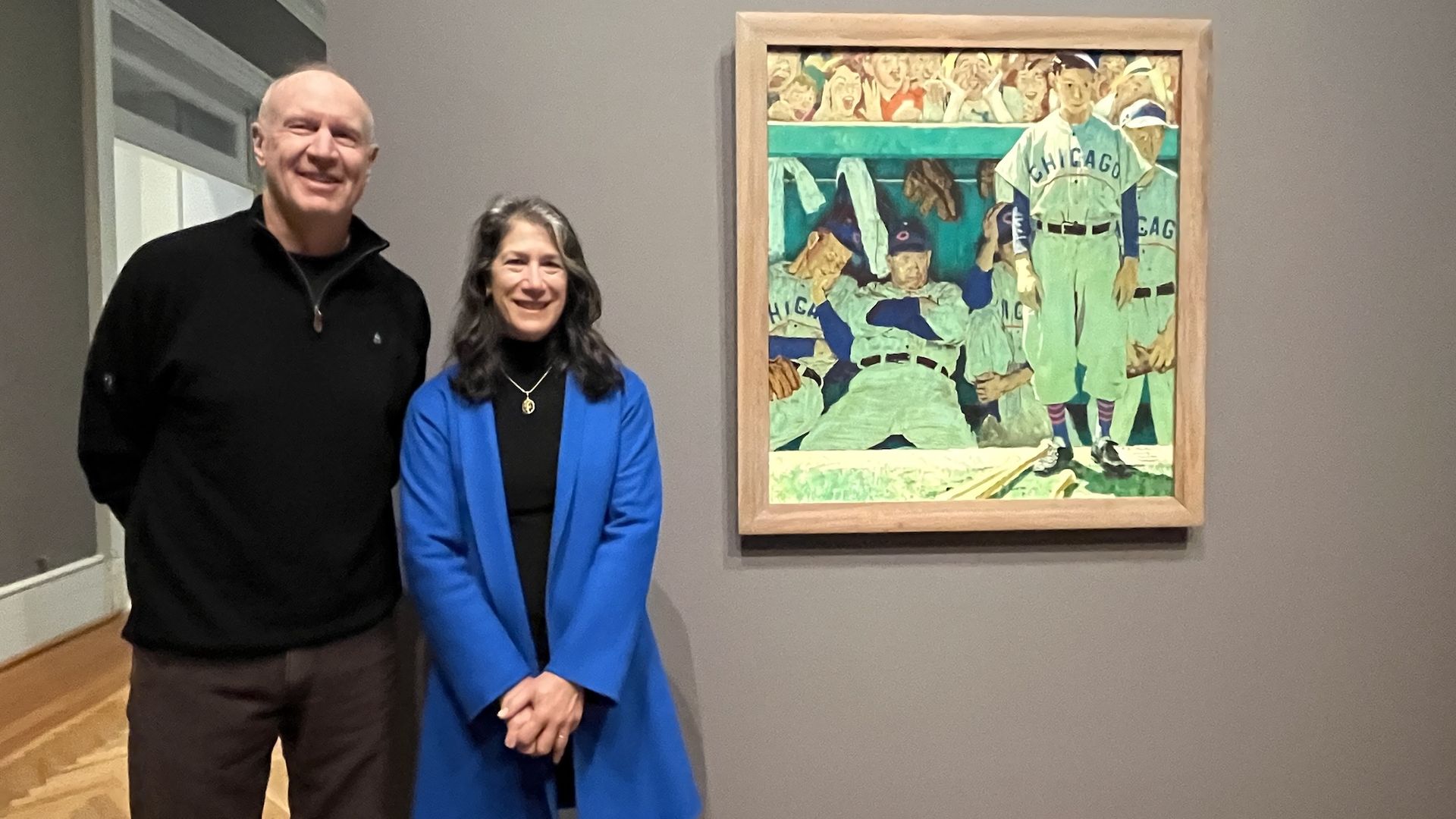 A man in a black sweater and a woman in a blue coat stand smiling beside a framed painting of Chicago baseball players in uniforms in a dugout, mounted on a gray wall.