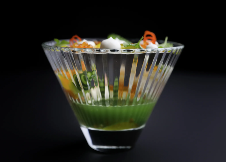 A small, elegant glass of king crab, cucumber, sudachi citrus and lemon balm in front of a black background.