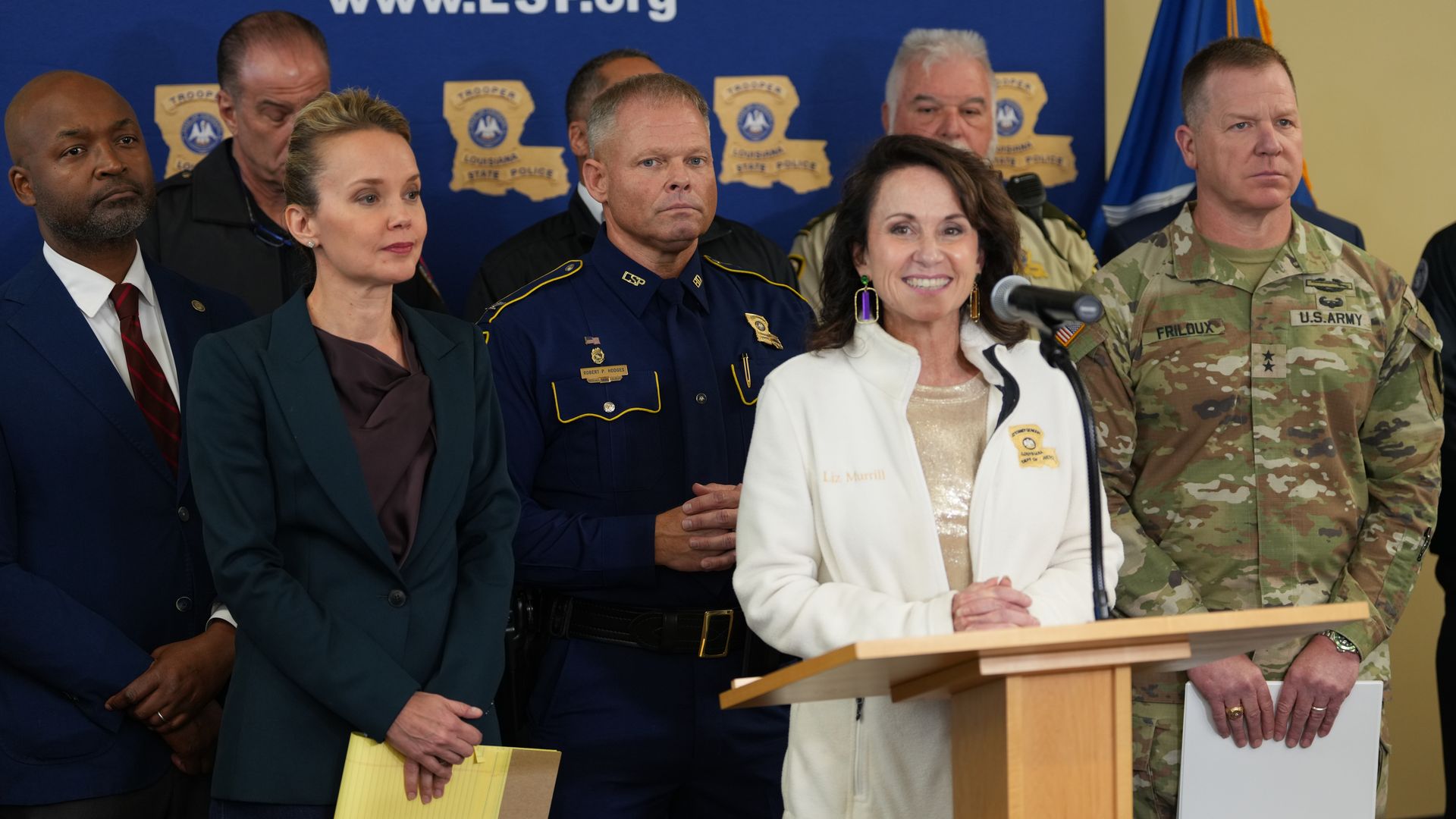 Group of officials at a press event: a uniformed police officer, a smiling woman at the microphone, a soldier in camo, and other officials against a blue backdrop with police patches and flags.