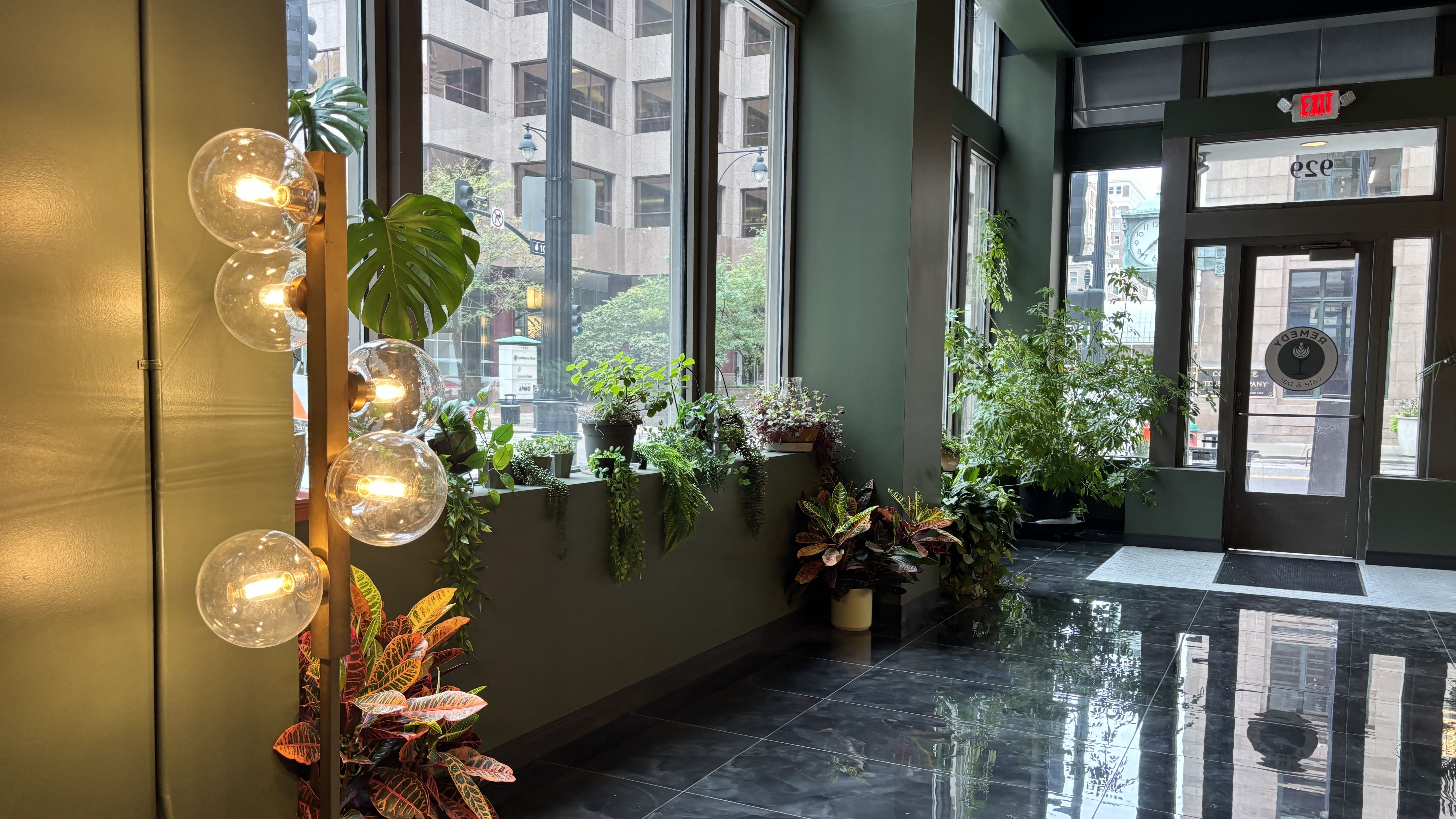 Modern lobby with dark green walls, large glass globe light fixtures on a gold stand, a row of potted plants along the window, glass door with EXIT sign, city street and clock outside, glossy black floor.