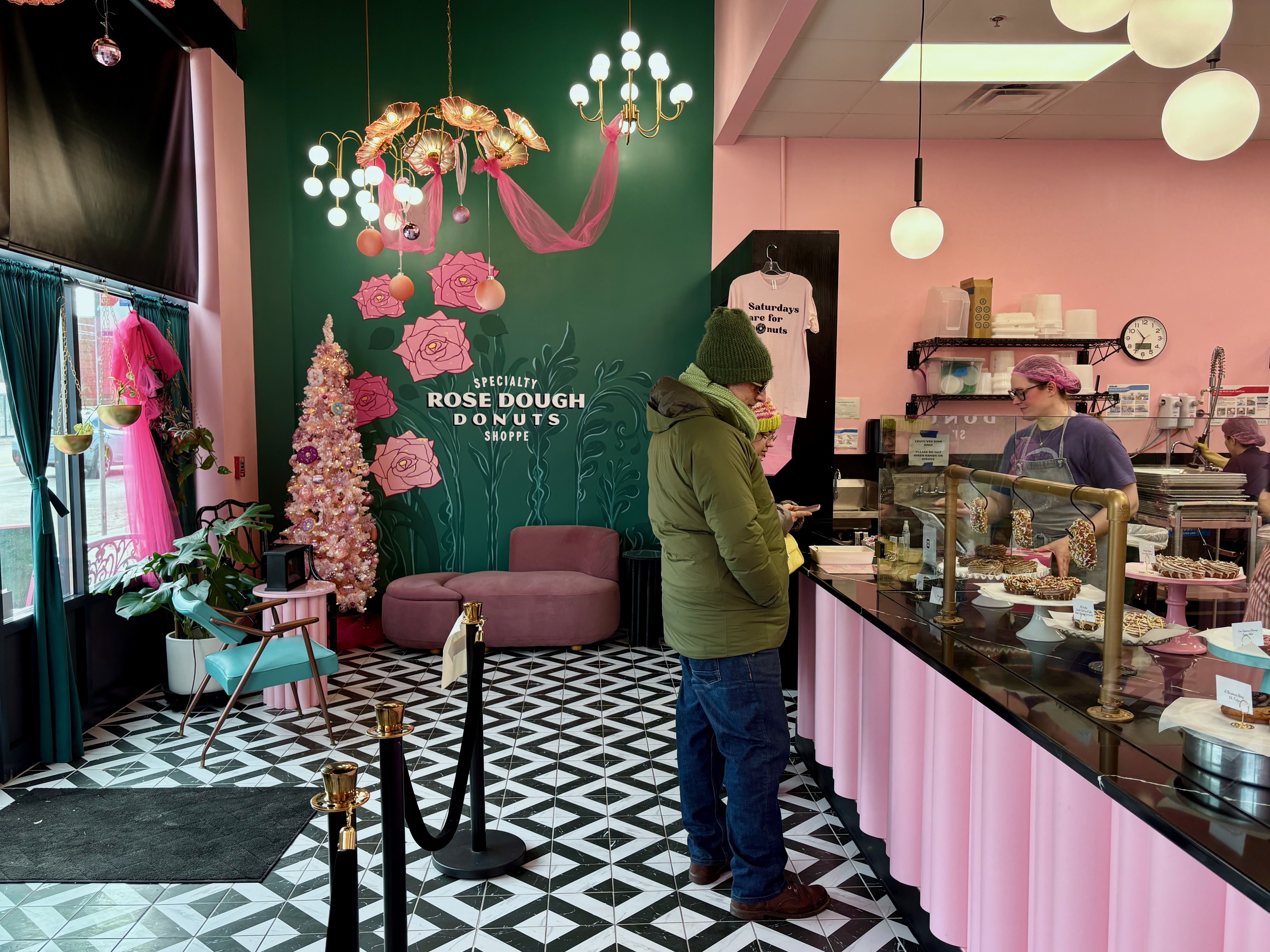Interior of Rose Dough Donuts shop with pink and green decor, pink Christmas tree, customers in winter coats at donut display counter, staff serving behind glass.