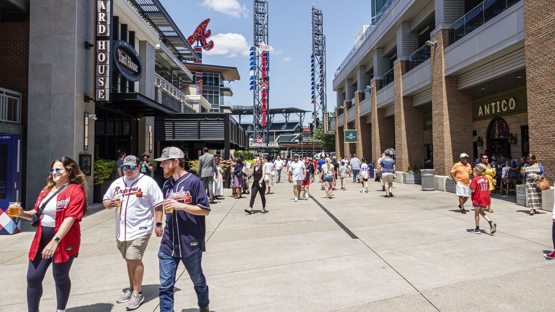 People wearing Atlanta Braves shirts and memorabilia and holding beers walk in a food plaza outside the team's stadium. A pizza place called Antico can be seen on the right. 