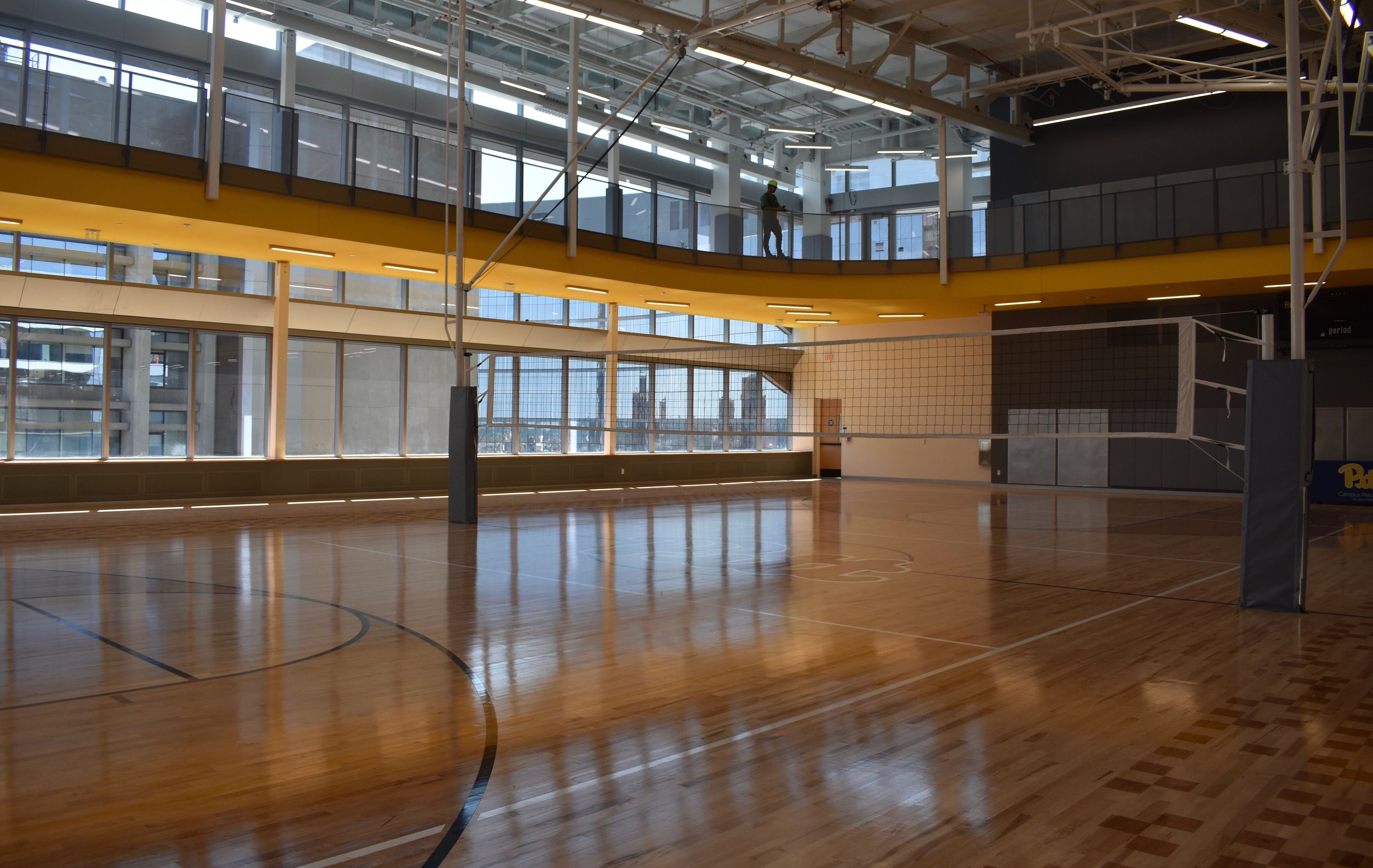 Empty indoor volleyball court with polished wood floor, black net, large windows, yellow ceiling accents, and a person standing on the upper level balcony.
